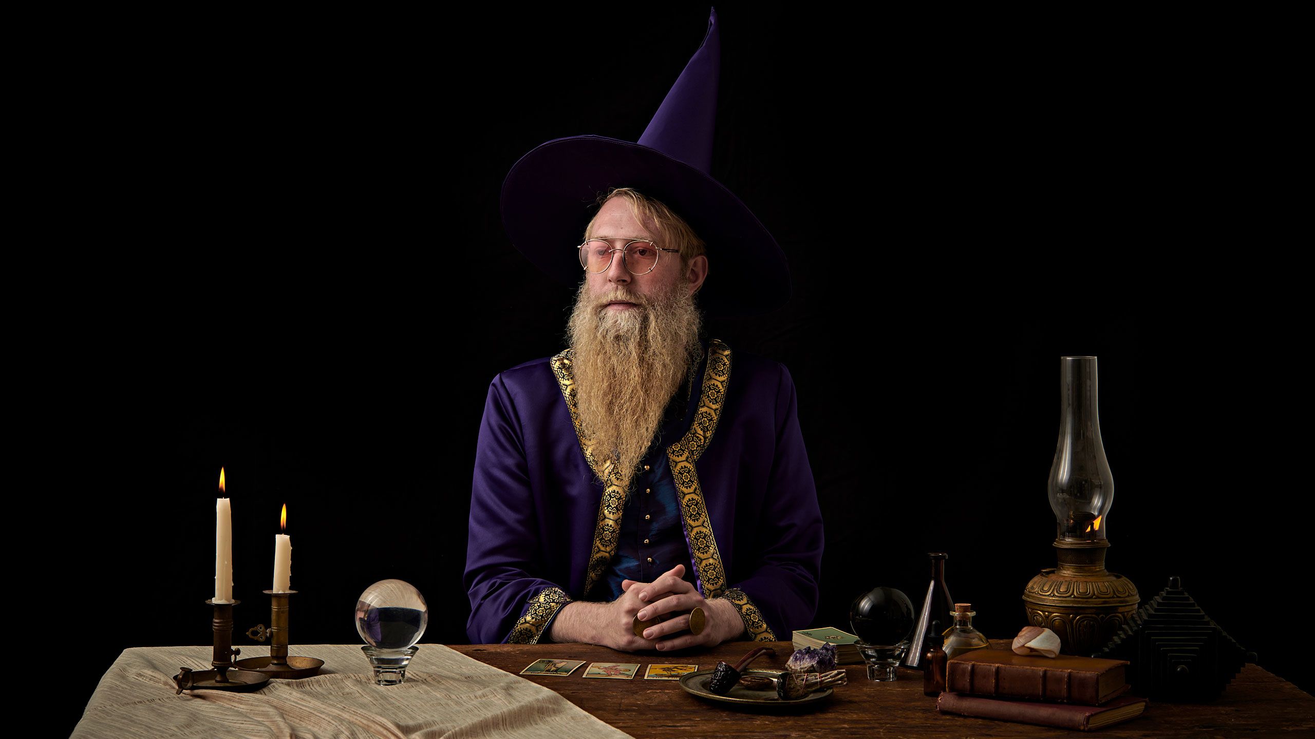 Devin Person sits and a table wearing purple robe with gold trim and a black wizard hat. Sitting on the table are two lit candles, a crystal ball, tarot cards, various potions, an old book and a kerosene lamp.