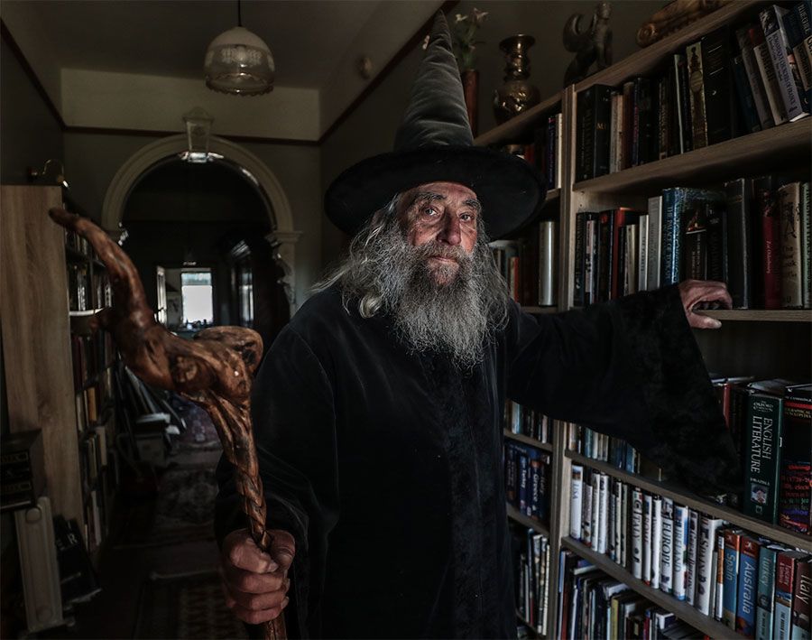 The Wizard of New Zealand standing the a hallway of an old house, leaning on a bookshelf full of books.