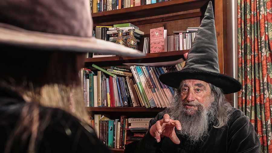 The Wizard of New Zealand gesticulates his hand in animated discussion with Ari while sitting together at the table.