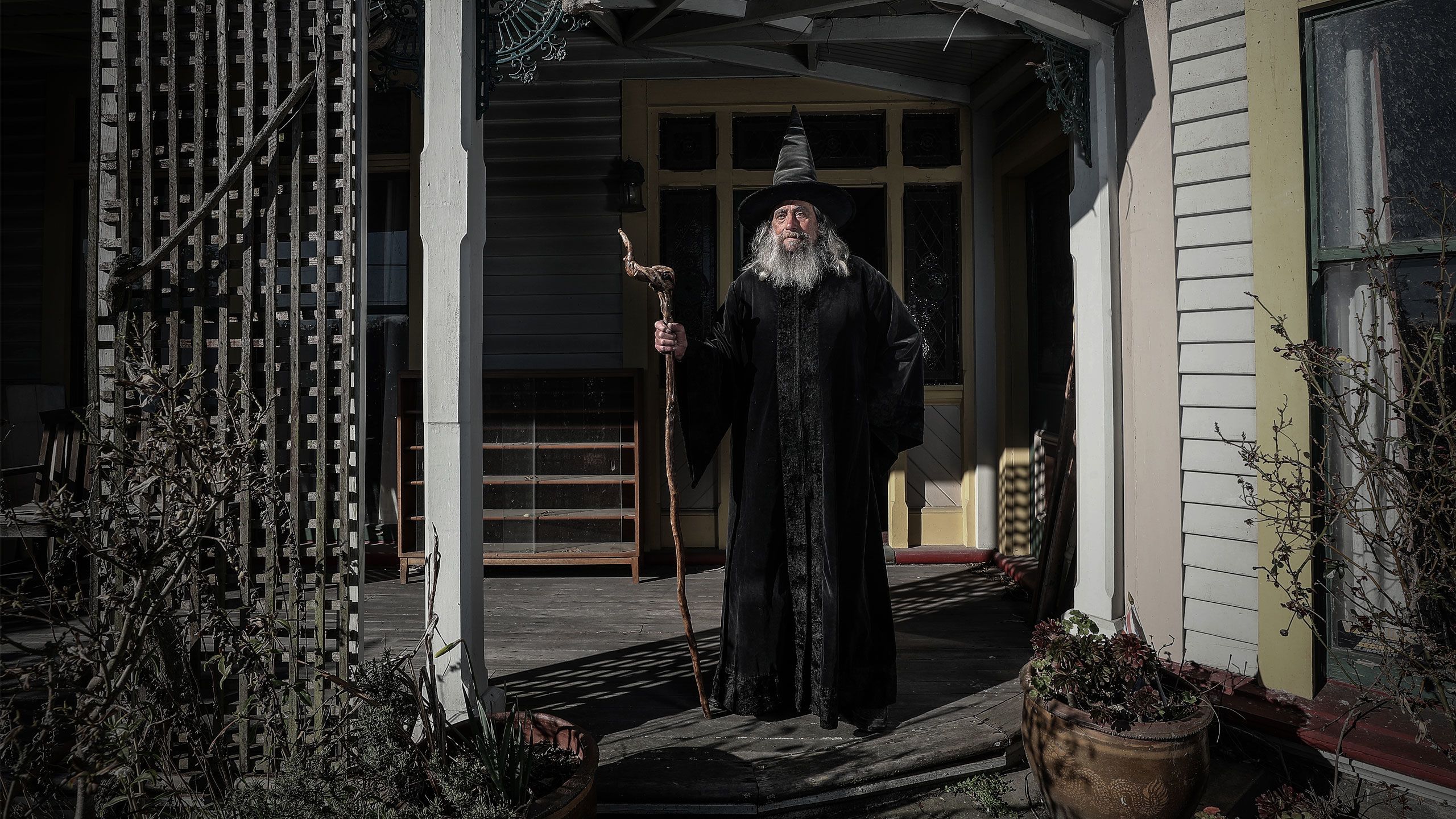 The Wizard of New Zealand standing with his staff in the porch of an old house.
