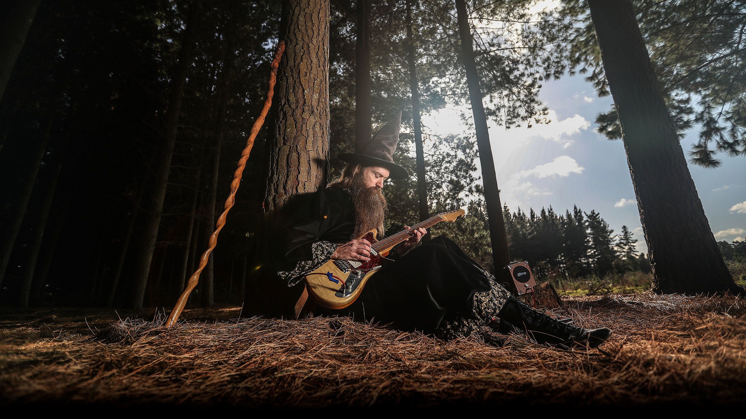 Ari sitting playing his guitar at the foot of a tree in a forest.