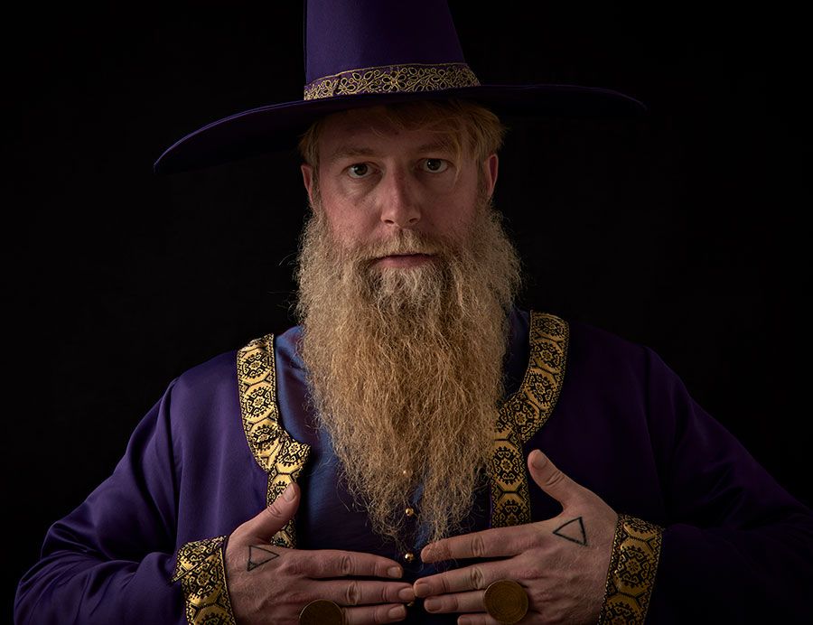 Close up of Devin who has raised his hands to his chest which reveal a large circular ring and a triangular tattoo on each hand.