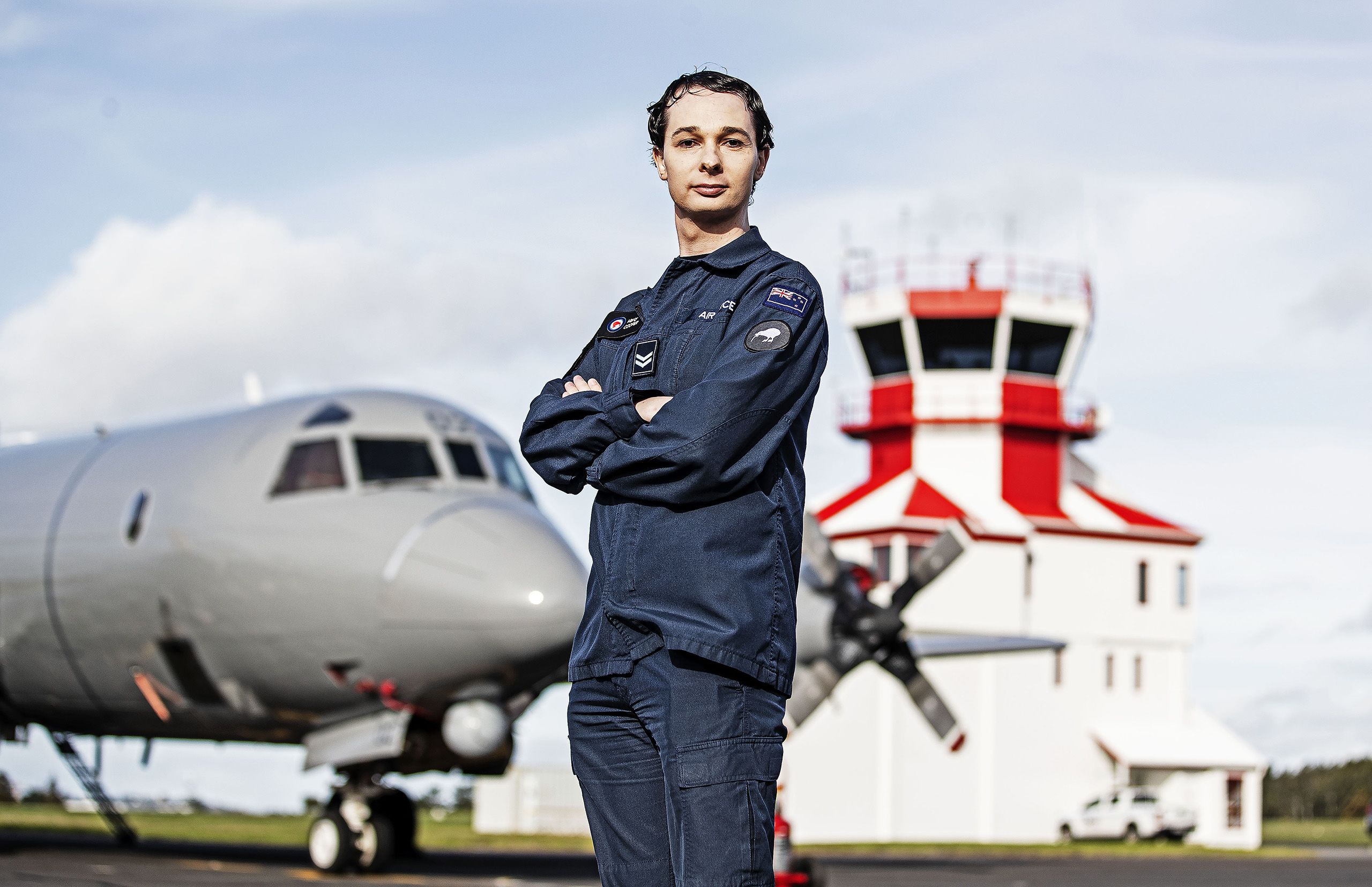 Marci stands in the foreground wearing her airforce uniform, arms folded with a military airplane and tower in the background.