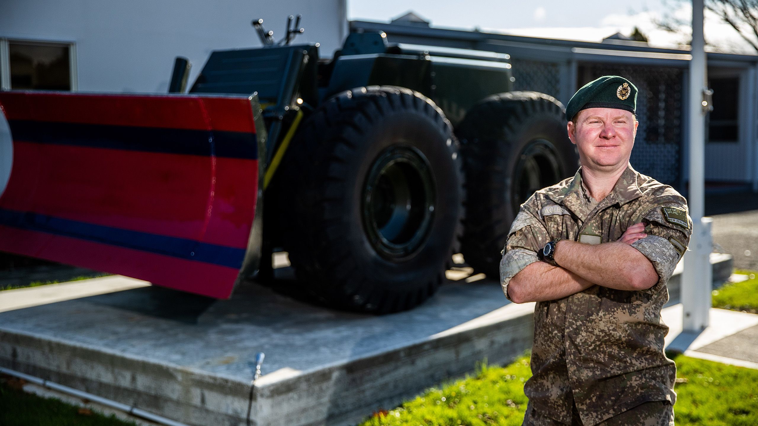 In army uniform, Tony stands in front of a military tractor with his arms folded.