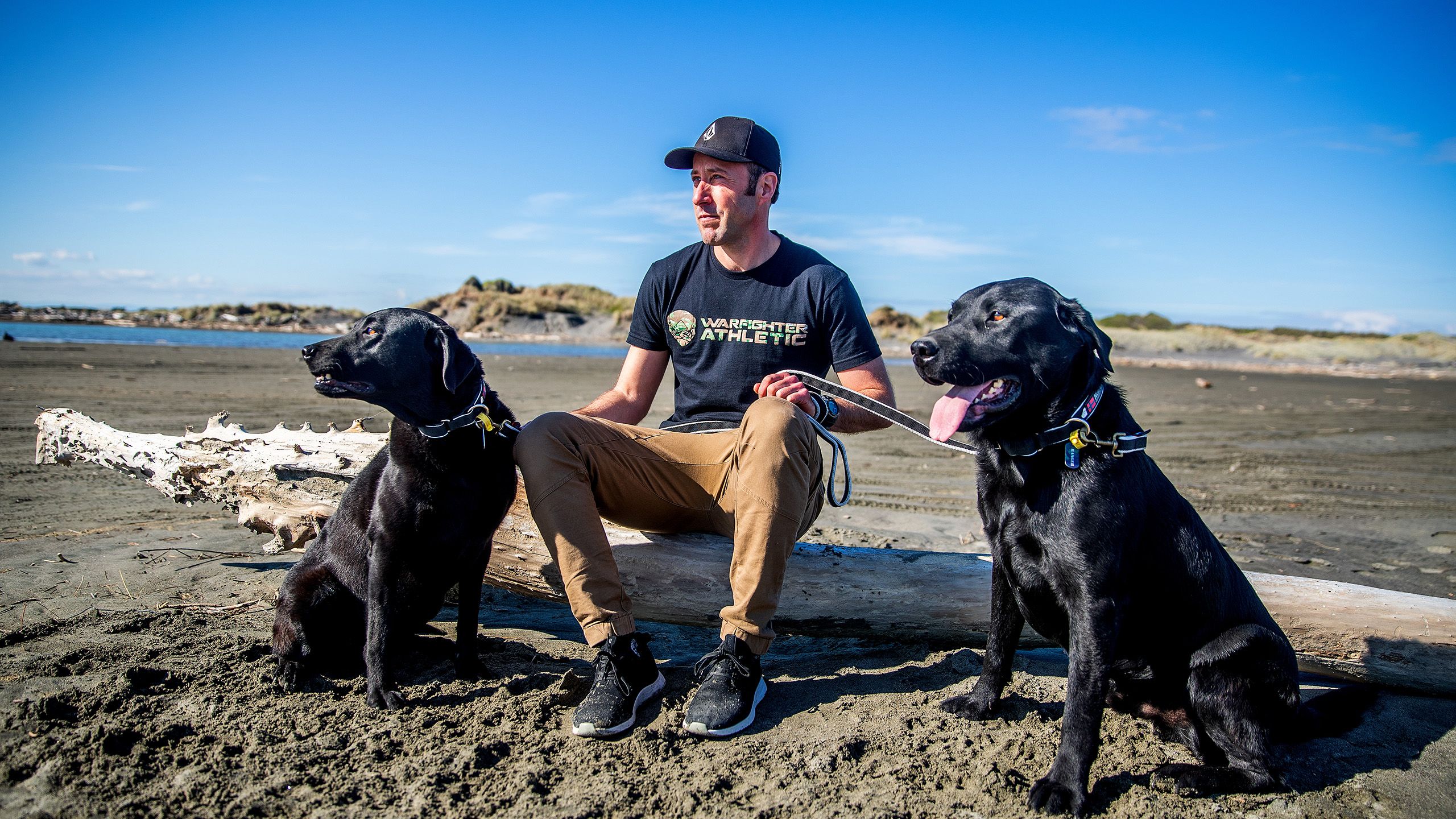 Mark sits on driftwood at the beach with his two black Labrador dogs.