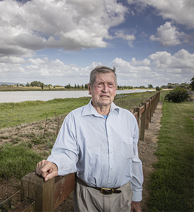 Peter Vandersloot rests his arm on a fence along the Waihou River which runs into the distance under a partly cloudy sky.
