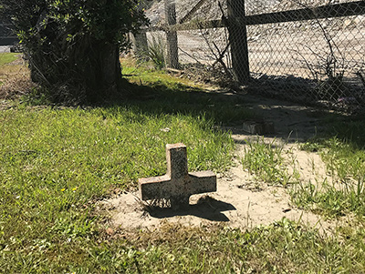 A stone cross sticks up from the ground in a dirt patch surrounded by grass along the side of a road. Behind an adjacent fence is a quarry.
