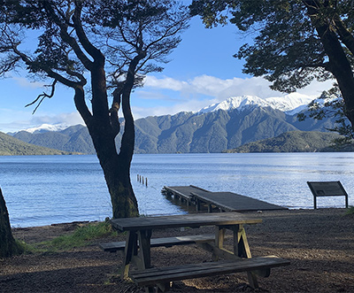 View of Lake Hauroko through a shaded area beneath trees. A dock extends into the lake. Snowcapped mountains line the horizon.