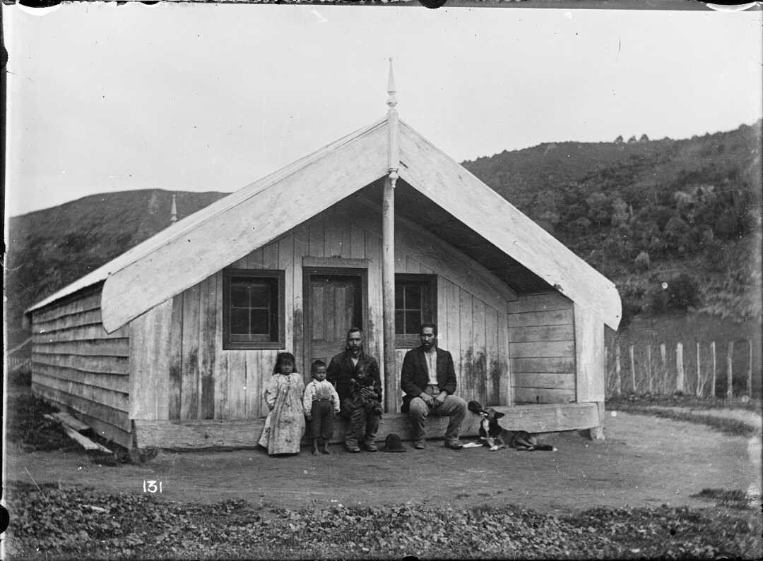 Two men sit beside children and dog in front of meeting house.