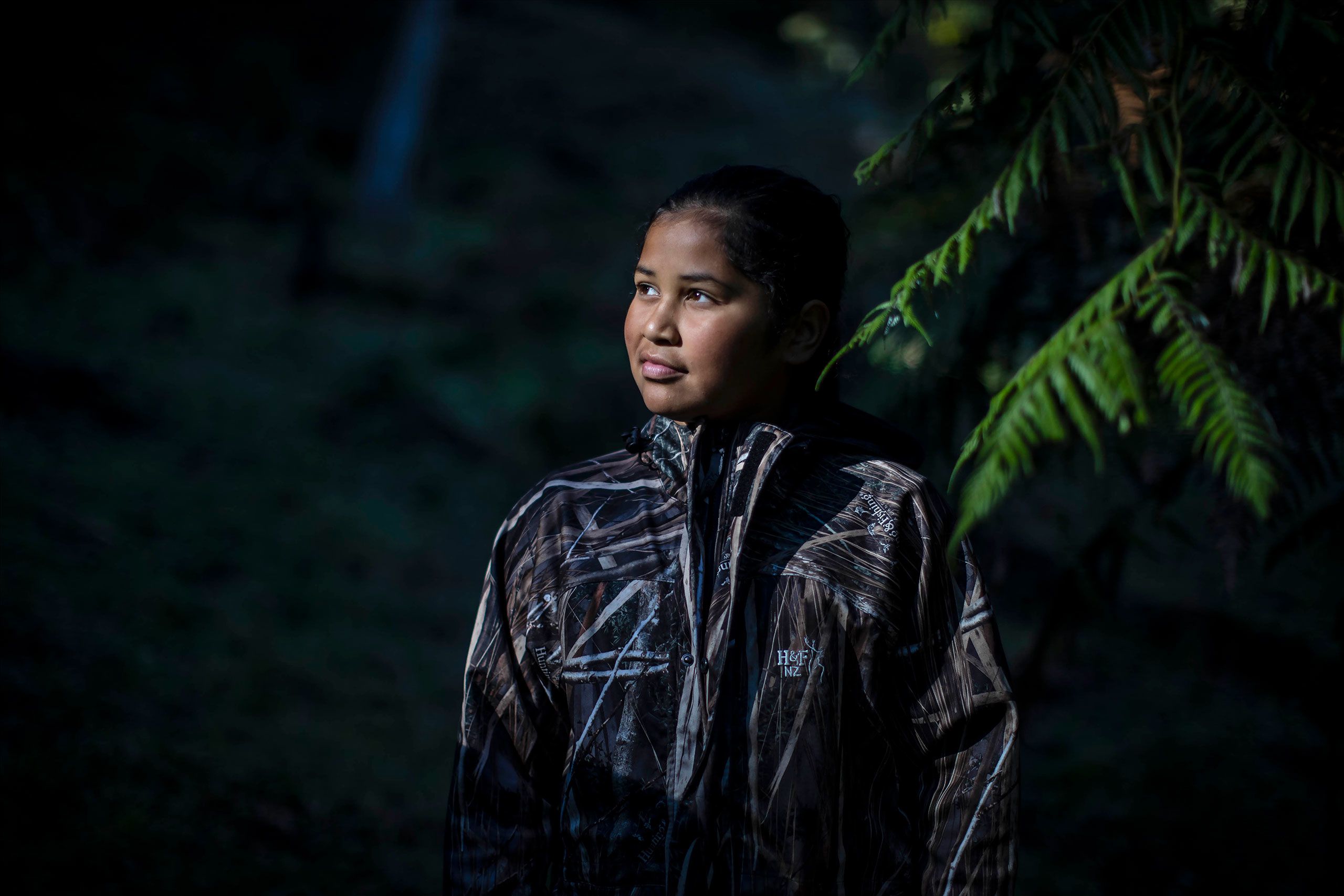 A dark background with the head and torso of Manu in the foreground. She looks up and to the left as the light shines on her face and some green fern fronds hang over her shoulder to the top and right.