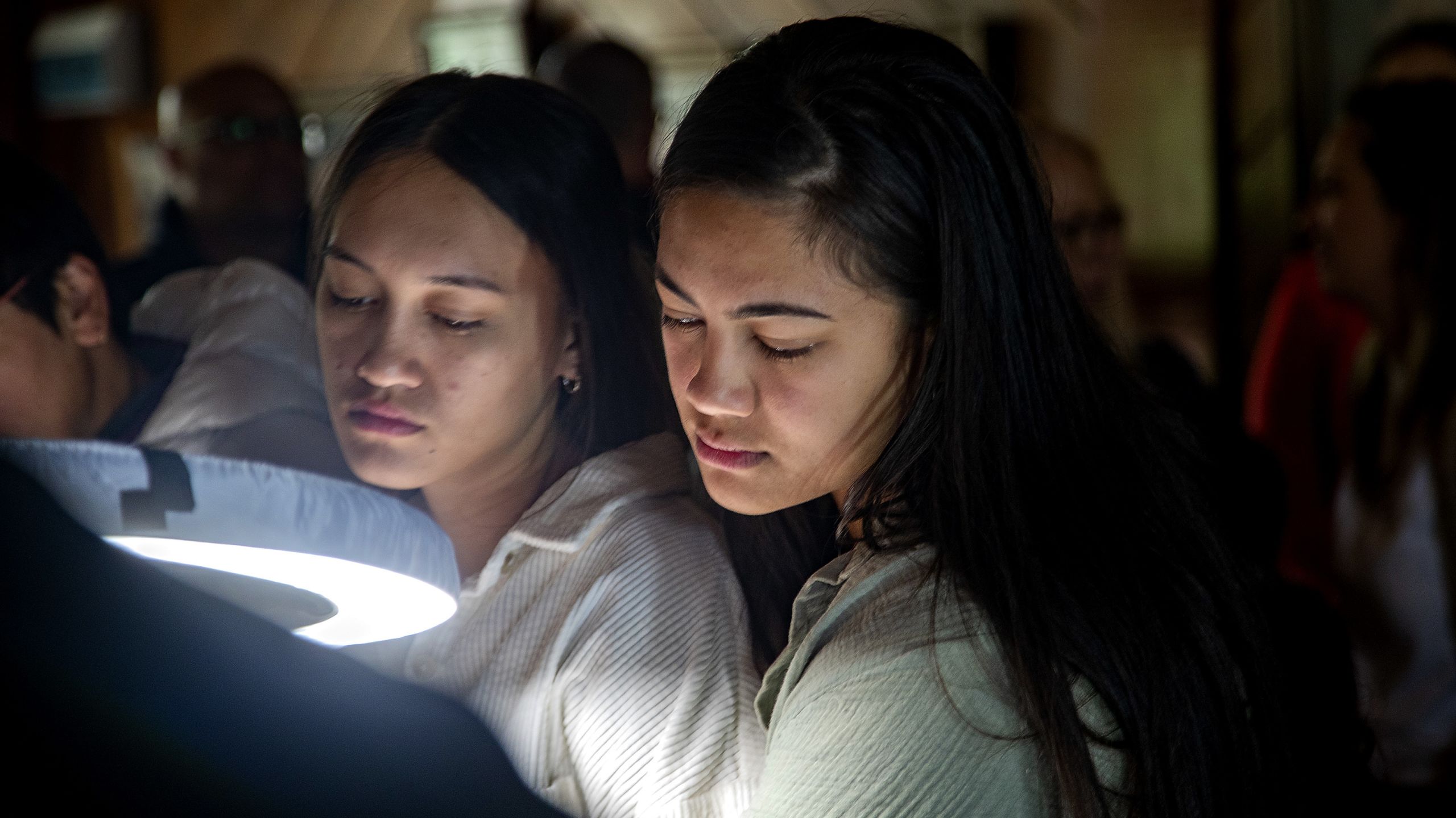 A close shot of Nuwyne's daughters, Hiria Mohi (left) and Aareta Mohi (right) watching the moko kauae process. They are standing next to the halo lamp used to light the room and are looking slightly downward, with solemn, focused expressions. Both have long, dark loose hair and are wearing light-coloured long-sleeved tops.