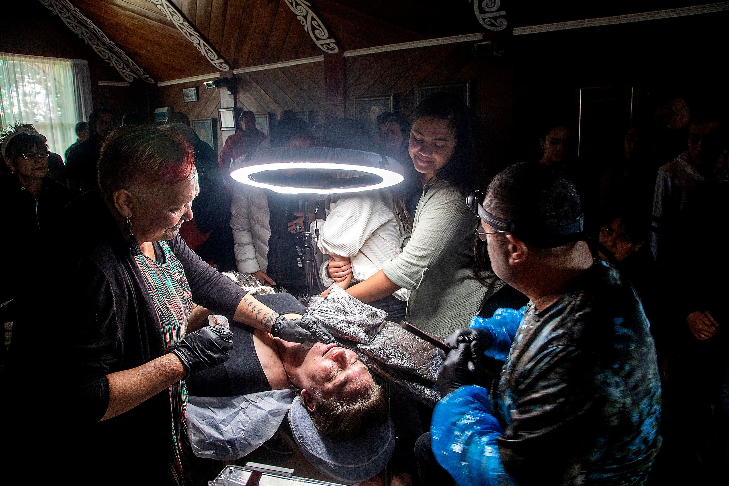 Nuwyne Te Awe Awe Mohi, lies on a tattooing table under a halo-shaped bright white lamp. She is dressed in a plain black top, with a woven, feathered cloak lying over her legs, and her eyes are closed. Her daughter, Aareta Mohi, is standing next to the table holding her arm for comfort and support. The tattooist, Hēmi Te Peeti, is at the head of the table, wearing black rubber gloves and a headlamp. His wife Takarea, an older woman with short dyed red hair, stands on the other side of the table and presses a soft cloth to Nuwyne’s chin to wipe away blood from the first chisel marks Hēmi has made. A crowd of people are dimly lit in the background. Everyone is inside an older building with a sharply pitched roof, wooden panelled walls and ceiling beams painted with traditional Māori designs in red and white.