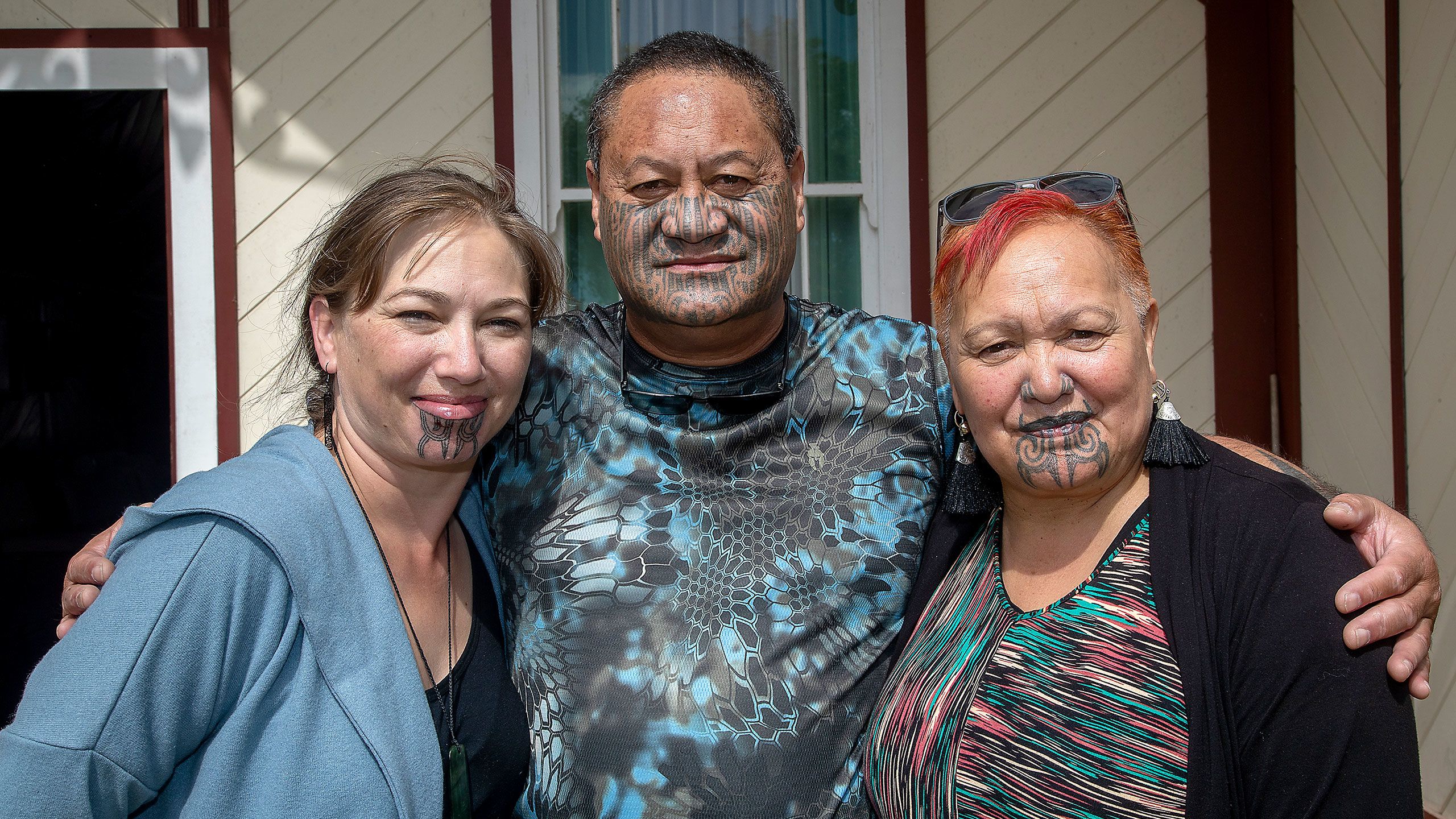 A waist-up shot of Nuwyne Te Awe Awe Mohi, Hēmi Te Peeti, and Takarea Te Peeti, outside Te Rangimārie meeting house, an older wooden building with cream-painted diagonally-laid weatherboards and dark red trim. Hēmi stands in the centre with his arms around the shoulders of the two women, staring directly into the camera. He wears a tā moko that covers his cheekbones to his chin and has close-cropped dark hair with specks of grey in it. He is wearing a blue and black patterned t-shirt and his glasses hang around his neck. Nuwyne is on the left, with he new moko kauae. The ink is dark and the lines are very distinct. Nuwyne has mid-brown hair pulled back into a plait and wears a pounamu pendant on a black cord round her neck. She is smiling with her mouth closed and squinting slightly in the sunlight. Takarea, Hēmi’s wife, is on the right. She has dyed bright red short hair and a moko kauae that extends to include her lips and the sides of her nose. She wears sunglasses on top of her head, tassled black earrings and has a multicoloured striped top on.