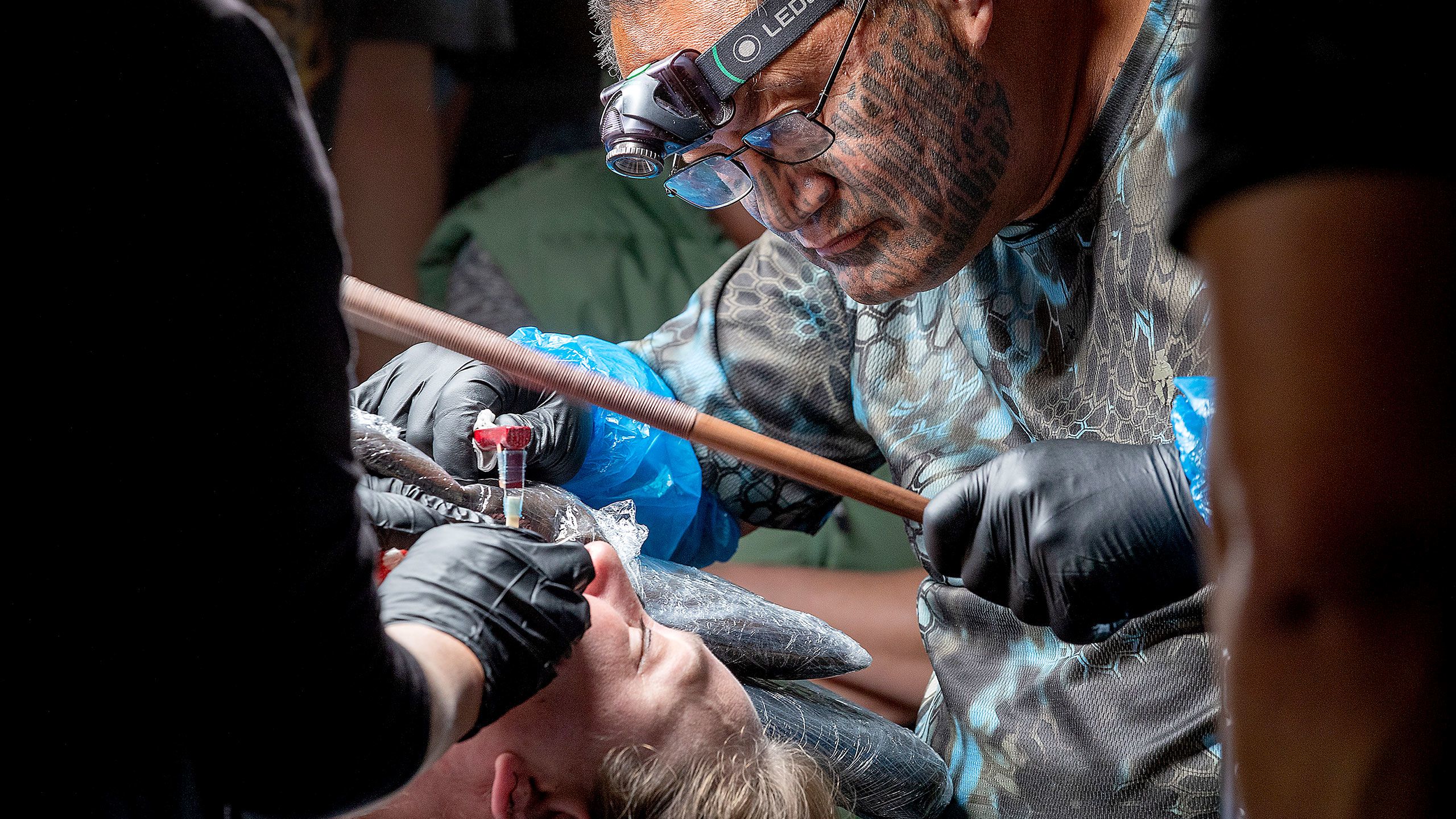 A close-up shot of the tattooist, Hēmi Te Peeti, leaning over Nuwyne Te Awe Awe Mohi as he begins chiselling her moko kauae. Hēmi wears glasses and a headlamp and has a tā moko (traditional Māori facial tattoo for men) covering his face from his cheekbones down to his chin. He holds his uhi (his traditional tattooing tool) in his right hand - the handle is made of red and clear plastic but the tip of the uhi is made of albatross bone. In his other hand he is holding a polished wooden stick that he uses to tap the top of the uhi to mark Nuwyne’s skin. The movement of the stick is slightly blurred in the image.
