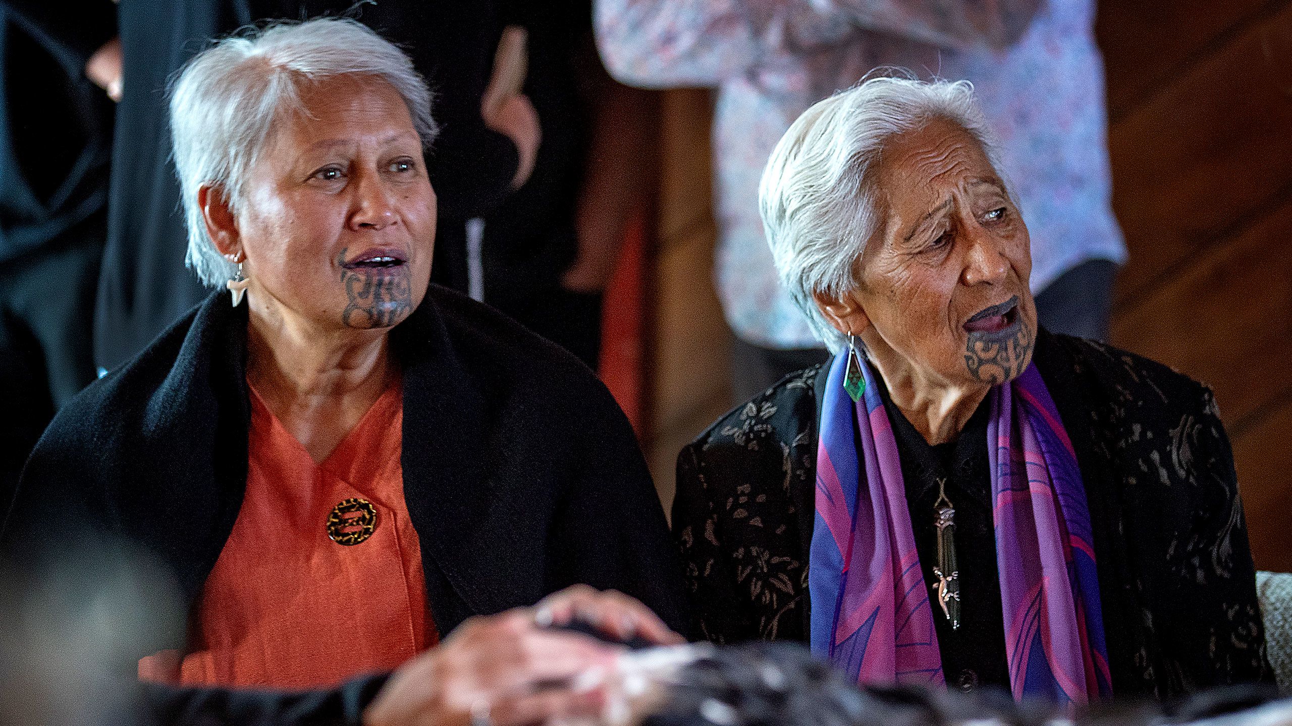 A close shot of two older Māori women in the room with Nuwyne Te Awe Awe Mohi. Both women have short, silvery hair and moko kauae. They have their mouths open and appear to be singing waiata. The woman on the right wears a salmon-coloured top with a brooch, a black blanket or coat covering her shoulders, and a shark’s tooth earring. The woman on the right, who appears to be slightly older, wears a long black top or dress with a bright pink and purple silk scarf around her neck. She wears pounamu earrings and a pounamu and silver pendant.