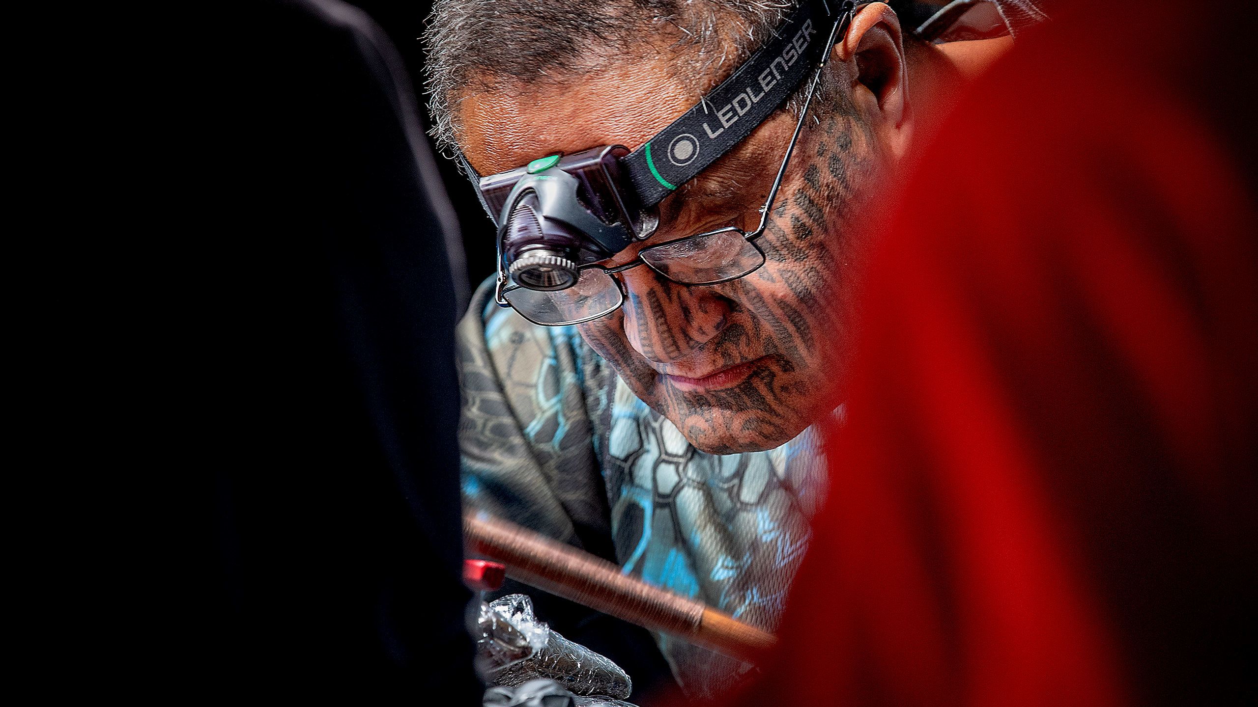 A very close shot of Hēmi Te Peeti as he continues tattooing. The photo shows just his face between the blurred shoulders of two people in the foreground. Hēmi wears glasses and a headlamp and has a tā moko (traditional Māori facial tattoo for men) covering his face from his cheekbones down to his chin. The wooden stick he holds to tap his uhi (traditional chiselling tool) is blurred, indicating he is moving it up and down rapidly.