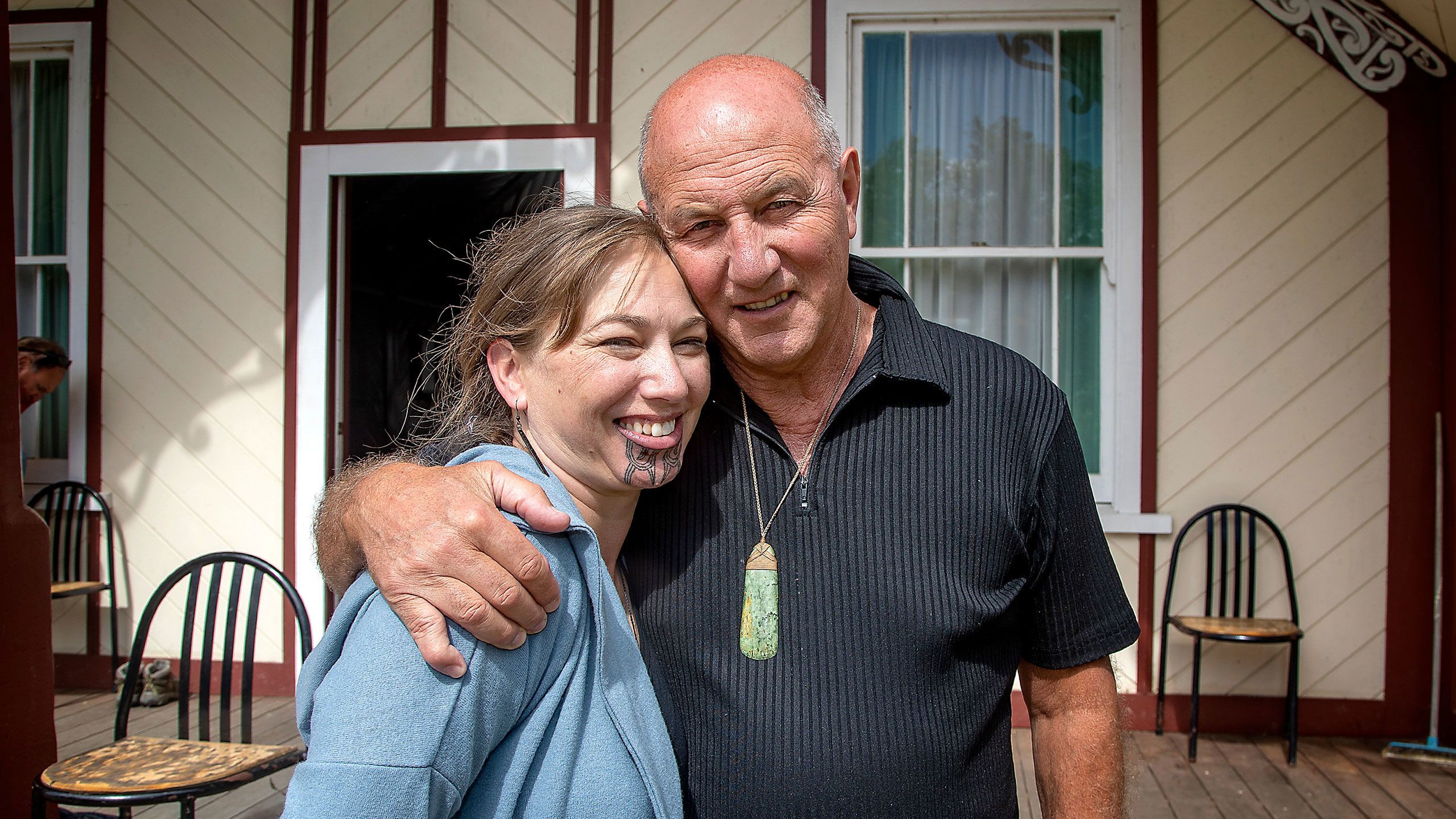 This photo is taken outside Te Rangimārie meeting house, an older wooden building with cream-painted diagonally-laid weatherboards and dark red trim. It shows Nuwyne Te Awe Awe Mohi and her father Wiremu Te Awe Awe, who has his arm protectively around his daughter and his cheek resting against her forehead. He is balding, and his forehead and eyes are creased. He wears a short-sleeved black top and a large pounamu toki pendant on a fine silver chain. Nuwyne wears a mid-blue hoodie and is smiling broadly with her mouth open. Her new moko kauae is dark, with distinct lines. Her chin is slightly red because the moko has only just been completed. They are lit by bright sunlight.