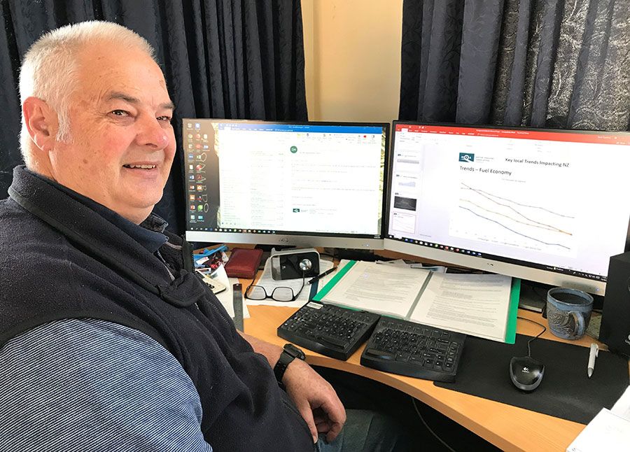 Motor Industry Association chief executive David Crawford is seated at a computer with two monitors. He is turning in his chair to look at the camera. He has very short grey hair and wears a dark zip-up vest over a blue shirt. One of the monitor screens shows a graph labelled 'Trends - fuel economy'. The line on the graph is trending down.