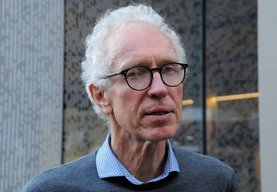 Alistair Woodward is photographed from the shoulders up. He is looking at the camera with a neutral expression. He is middle-aged with white, slightly curly hair and wears dark-rimmed glasses, a blue-checked shirt with a grey jersey over top.