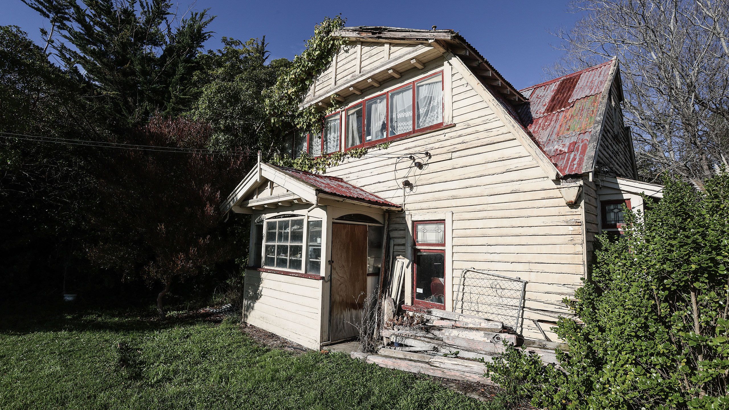 The front of Ferrymead House. The cream paint is weathered and the red paint has nearly rusted off the corrugated iron roof. Creepers are growing up the house and have overtaken the upper lefthand corner of the building. An entranceway to the covered porch has been covered up with plywood and a jumble of old timber and metal fencing sits in a pile next to the house.