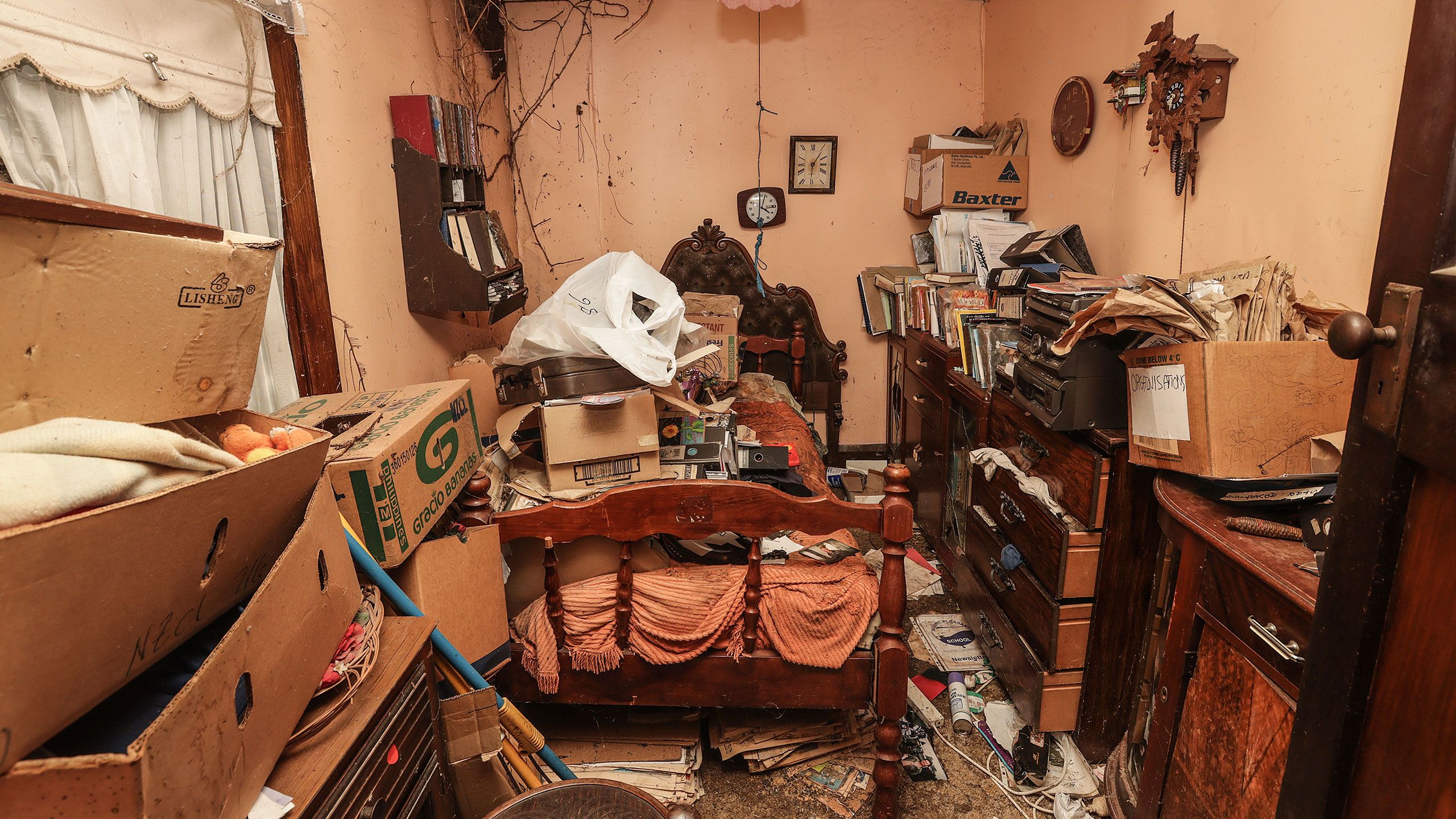 Philip Wright's old bedroom in Ferrymead house. A bed with wooden bed ends is piled with cardboard boxes, ring binders, and bags of belongings. More cardboard boxes are stacked high in the foreground. Several chests of wooden drawers on the right are visibly full of clothing, which is overflowing from unclosed drawers. Two clocks hang above the bed on peach-coloured walls. There is barely any floor space visible under all the objects and junk in the room.