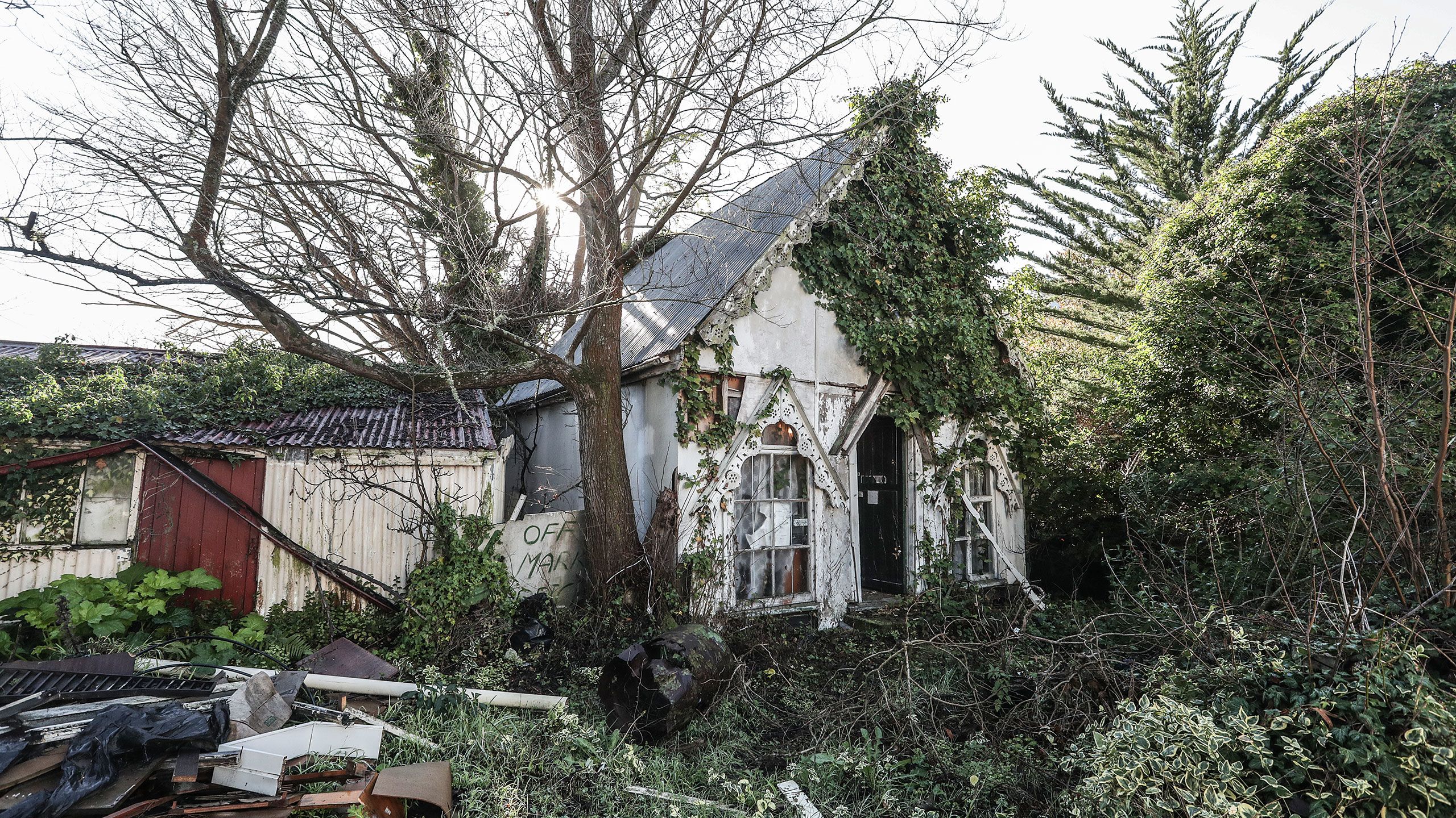A chapel-like building, which Philip Wright used to house objects and photographs related to the history of Ferrymead. The building is painted white with a grey iron roof and there is intricate wooden lacing adorning the eaves of the building and two dormer windows at ground level. The building is in a state of disrepair and creepers have overtaken one entire side. A newer-looking wooden out-building is attached to one side of the chapel but that is also overgrown with creepers.
