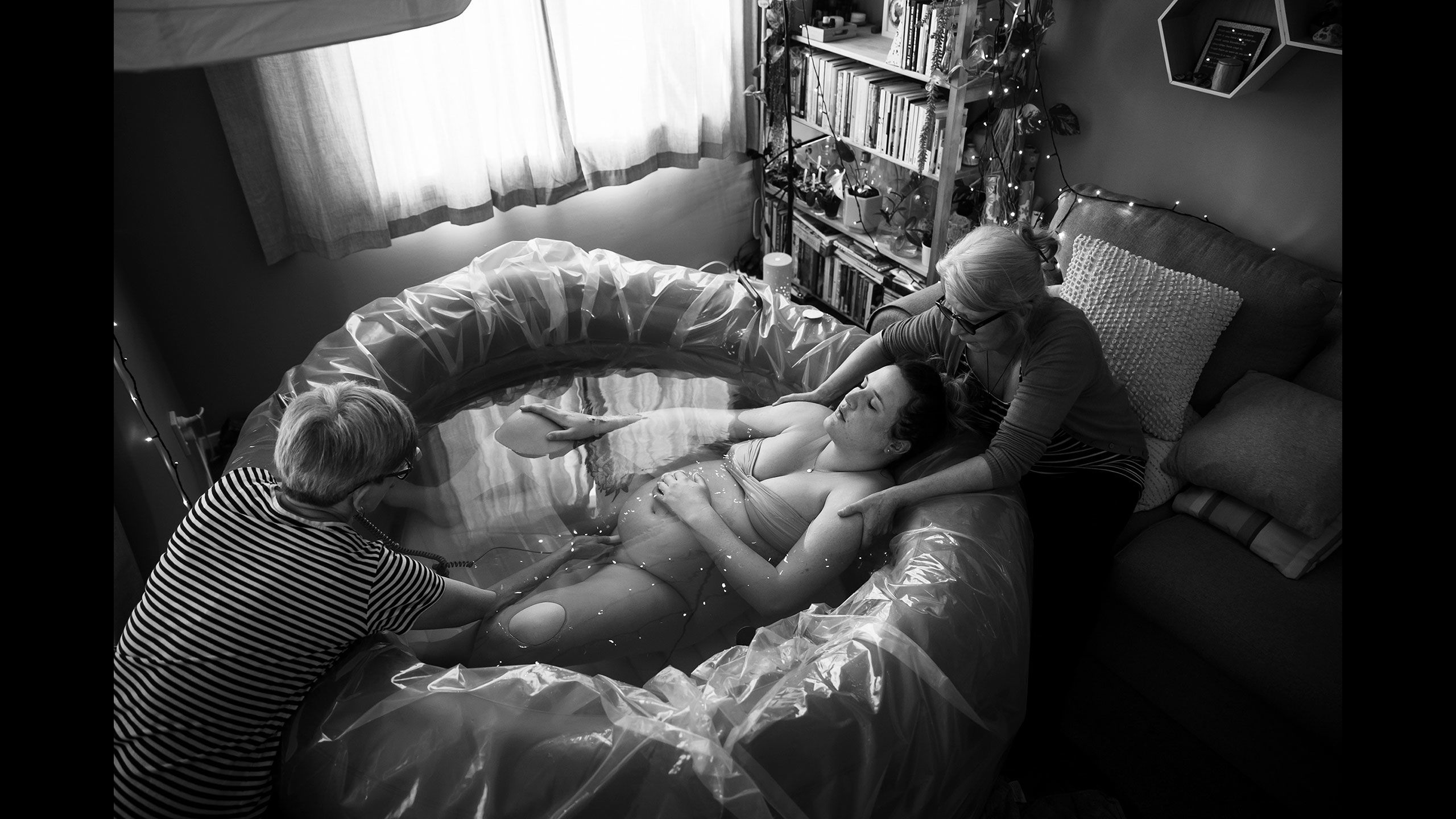 Jessica lies relaxed face up in the birthing pool with her hand rested on her baby bump. Her mother kneels behind her gently holding onto her shoulders. The midwife is checking the baby's heartbeat by holding a monitor onto the bottom of Jessica's belly.