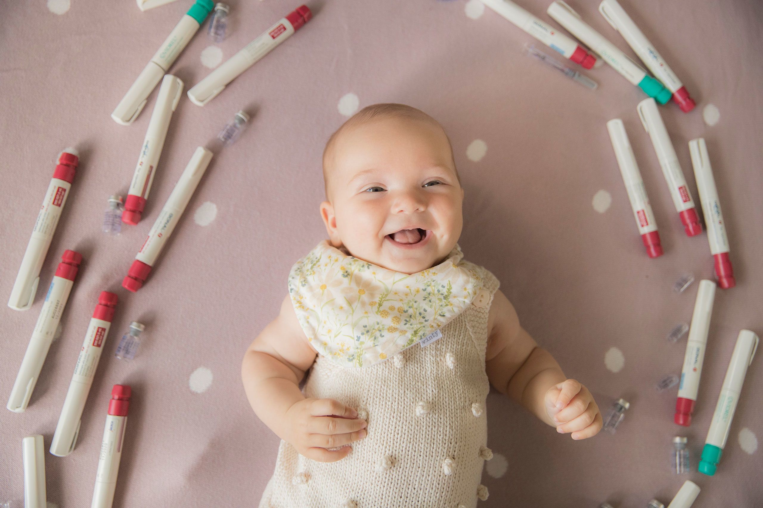 Looking down on Hazel who is giggling while lying on a dusky pink coloured blanket with a cream dot pattern. She is surrounded by the IVF home use equipment which helped to make her.