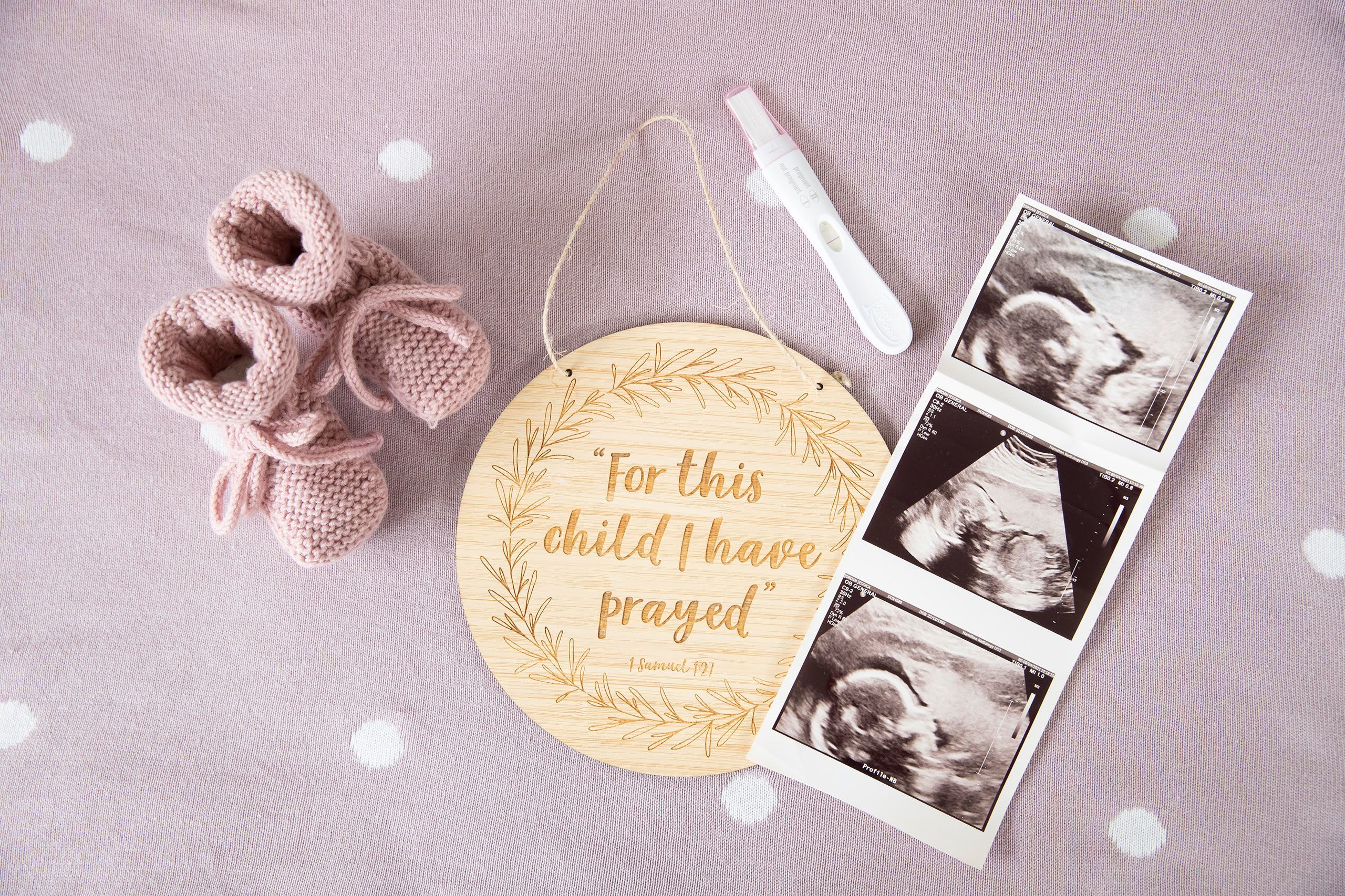 A scene of several items placed onto a pale pink blanket with white polka dots. In the middle is a circular wooden plaque with the words "For this child I have prayed" etched into it. Top left are a pair of dusky pink booties. Top right is the positive pregnancy test. Direct left is a strip of the ultrasound images of the baby in Jessica's womb.