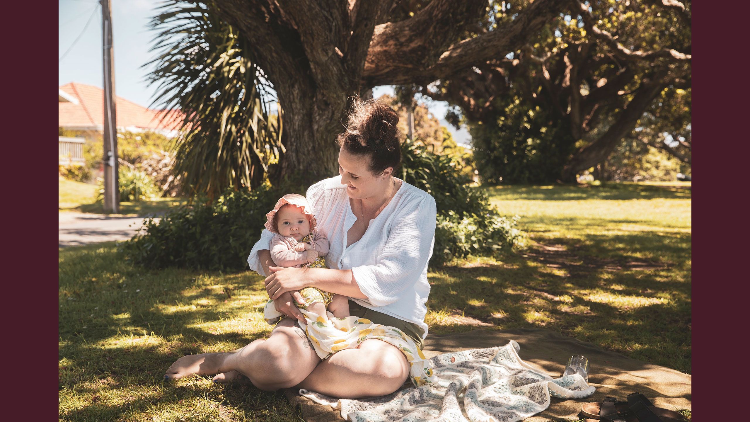 Jessica outside sitting under a tree holding Hazel in her arms. hazel is wearing a little bonnet to keep the sun off her face.