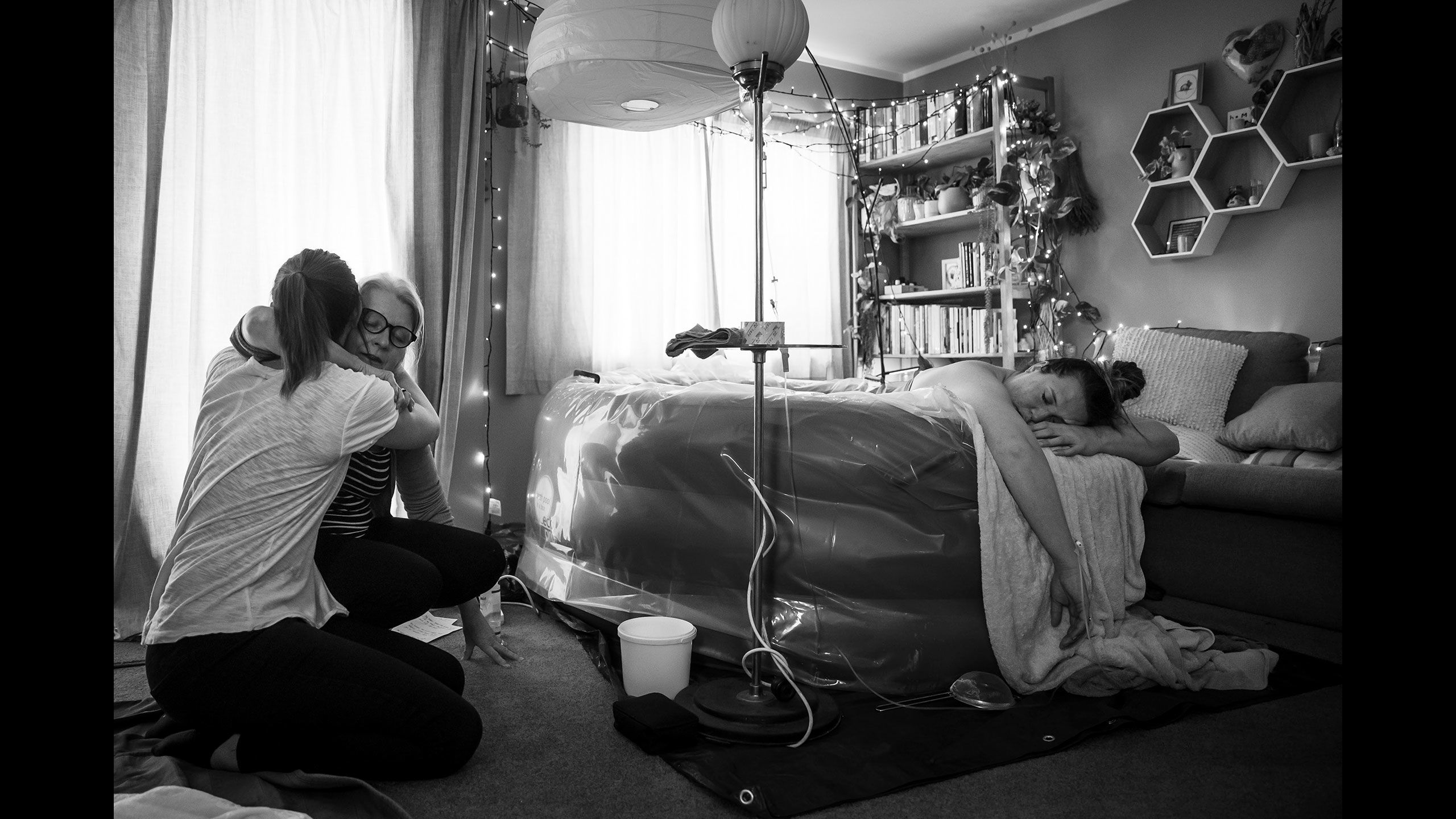 A black and white image of Jessica in her front room lying in the birthing pool on her front with her arms and head hanging over the edge of the pool. To the right, her mother and sister-in-law are hugging each other during a moment in between Jessica's contractions.