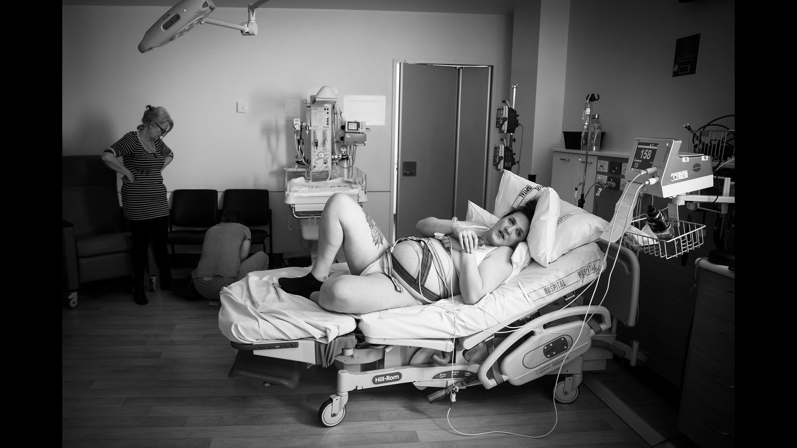 Black and white image of Jessica lying on a hospital bed connected to multiple tubes and lines. Her mother in stands in the background in attendance.
