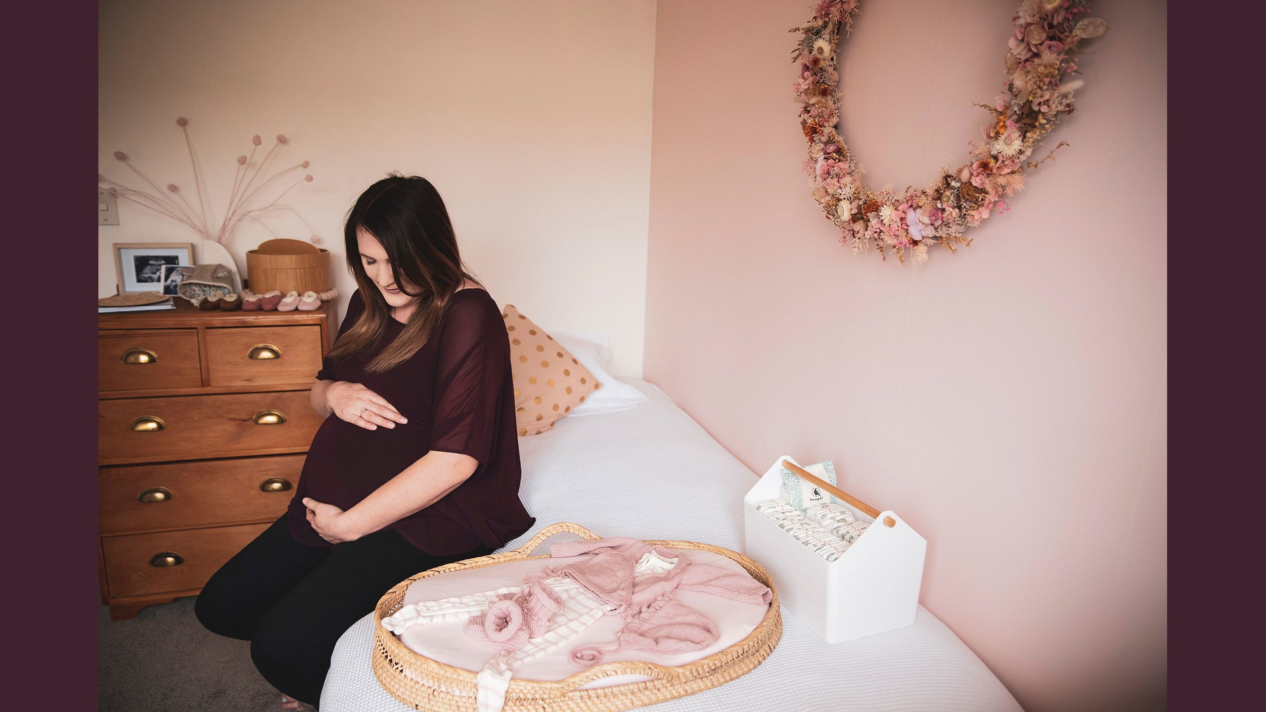 Jessica sits on a single bed in the nursery holding her baby bump. On top of the bed is a wicker tray covered in pink and white baby clothes.