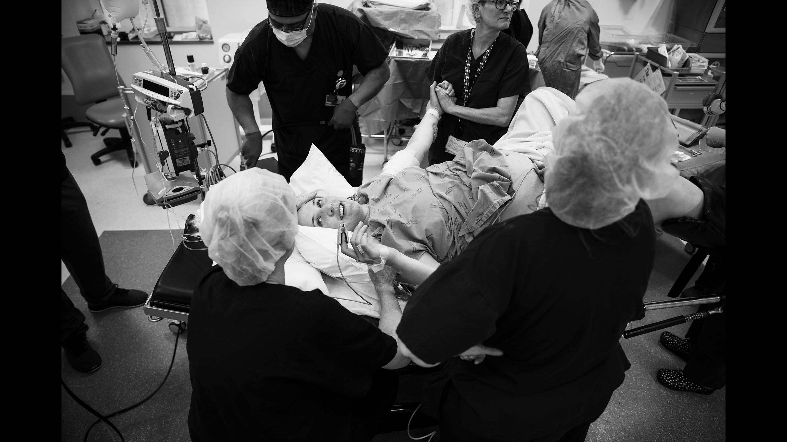 Jessica on lying on a hospital bed surround by pre-op staff preparing her for surgery. She holds onto her mother's hand who is by her side.