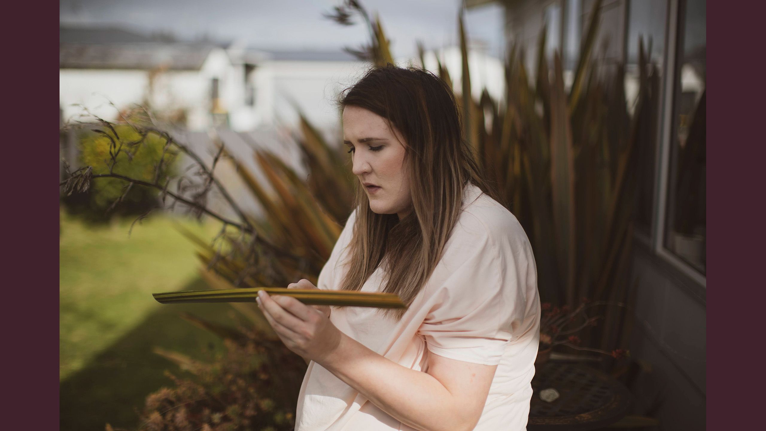 Jessica outside holding a piece of harakeke (New Zealand flax) in her hands preparing it for weaving. Behind her in the background is a large harakeke bush.