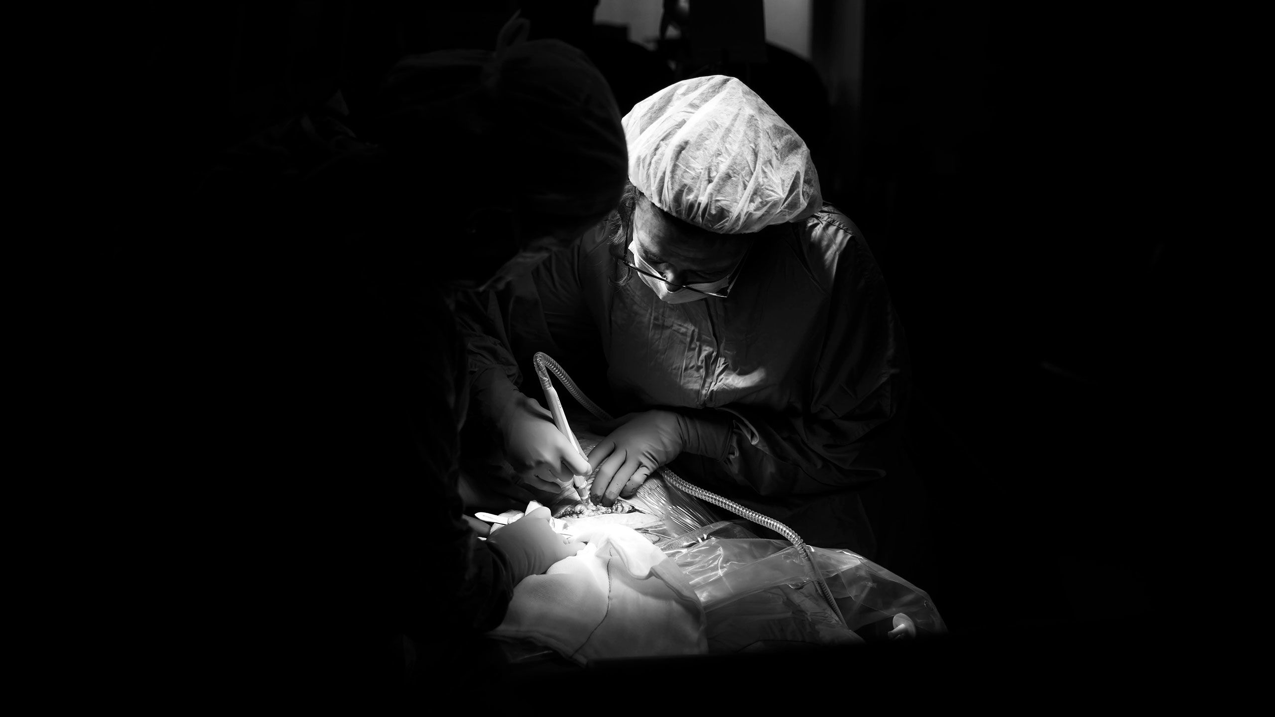 A stark black and white image of the surgeon starting the caesarean section procedure, by cutting into the skin of Jessica's belly. It is dark, with a spotlight on the surgeon and Jessica's belly.