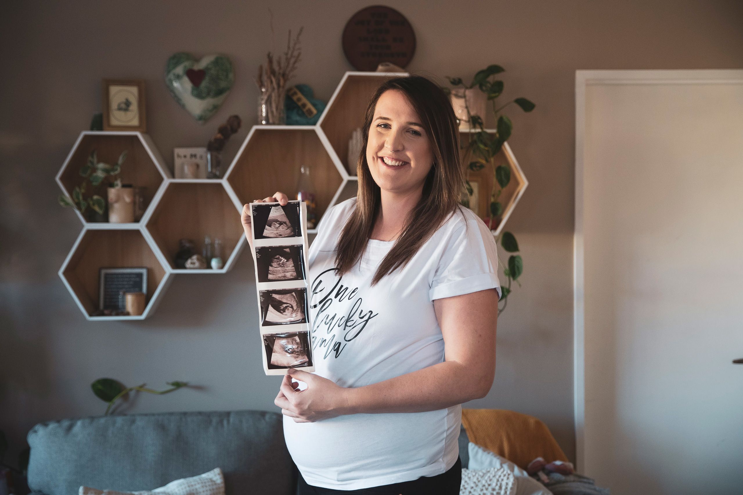 Jessica standing in her living room, smiling broadly and holding the strip of ultrasound images.