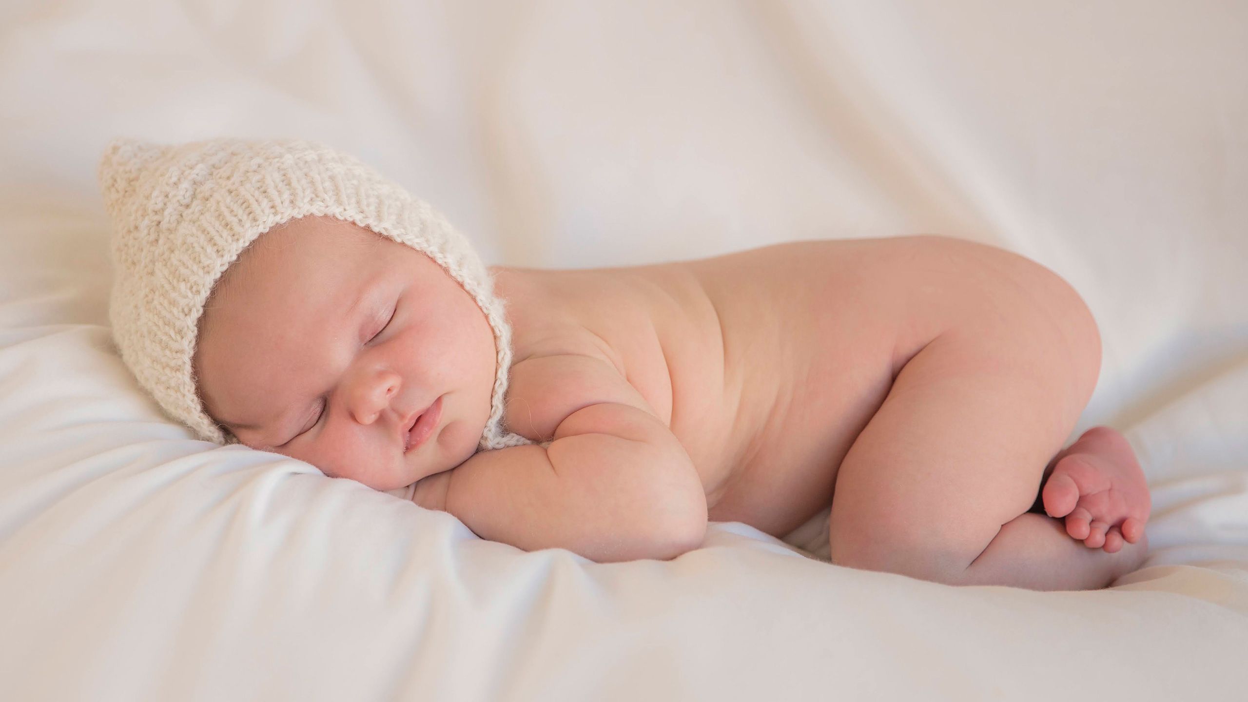 A cute soft focus image of Hazel sleeping on her front with her hands under her chin and her head sideways to the front. She is in her birthday suit wearing only a cream, woollen knitted bonnet.