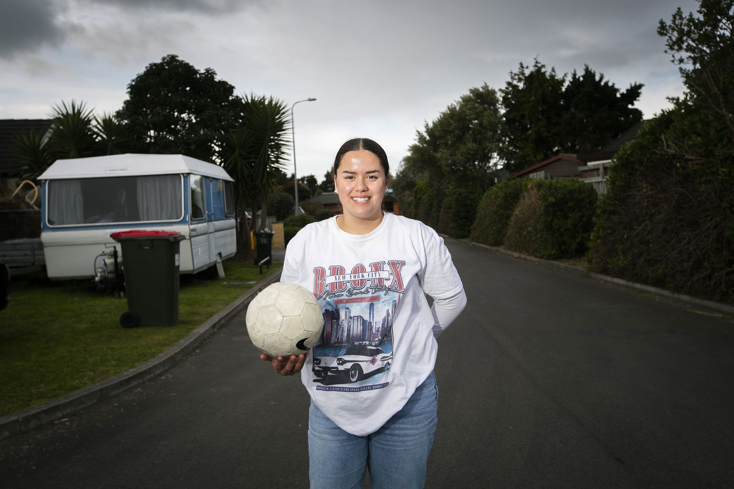 Brooke standing on the road near their home holding a soccer ball in one hand. She wears a Bronx New York City, long sleeved T-shirt and pale blue jeans.