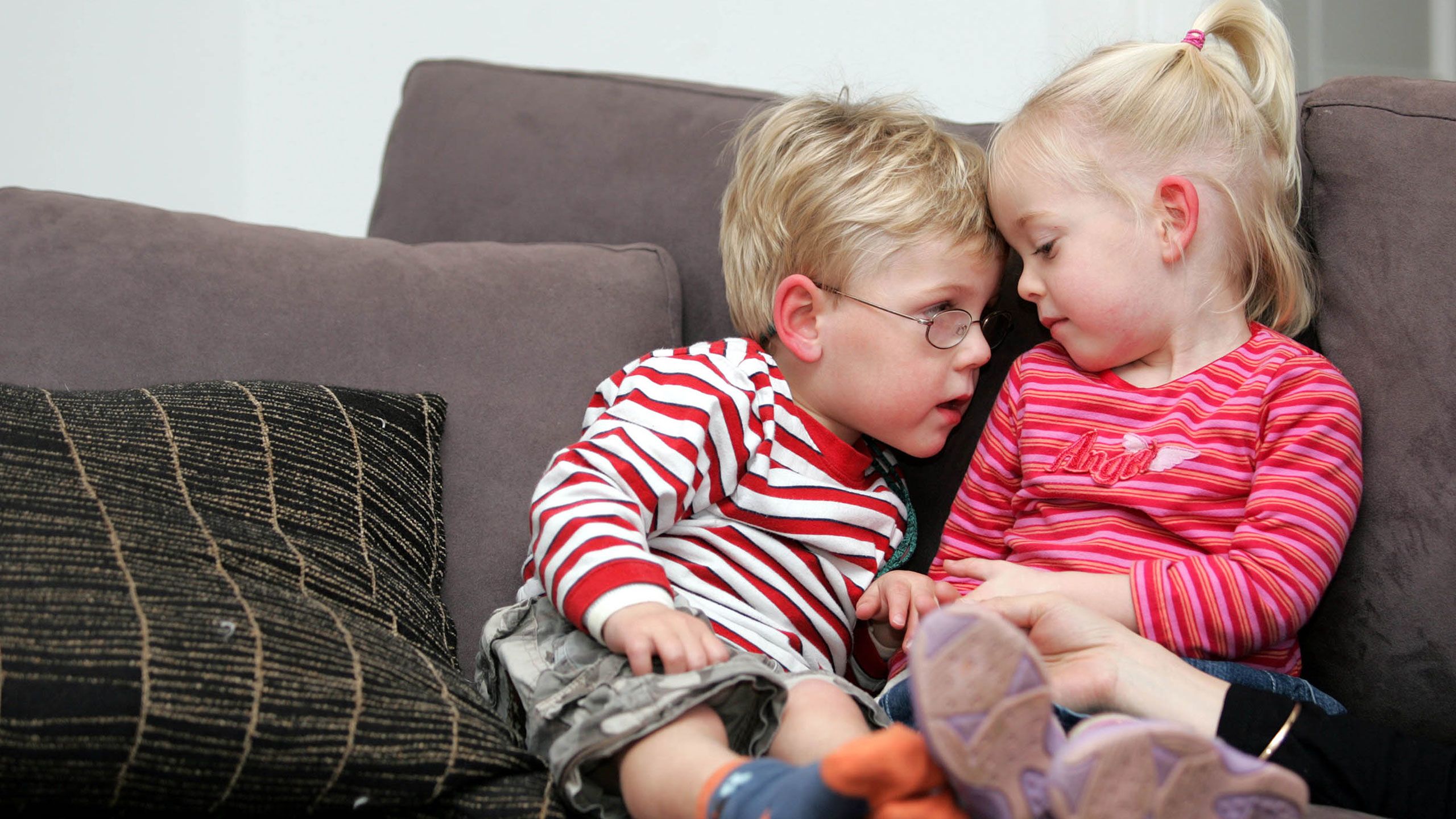 At three years old, Mac sits on a sofa next to his twin sister India. Mac is nuzzling up to her as she looks gently down on him.