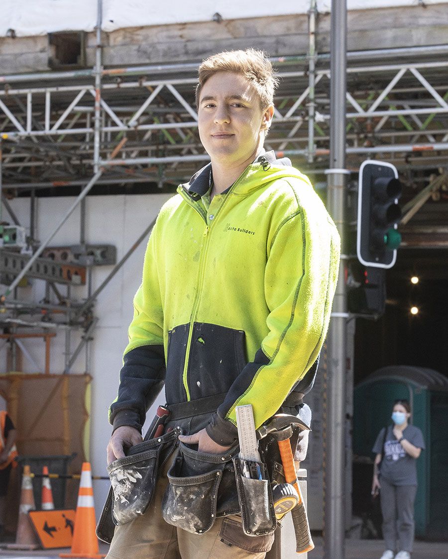 Cameron stands outside the building site in his high-vis sweatshirt. Around his hips he wears his tools in a holster.