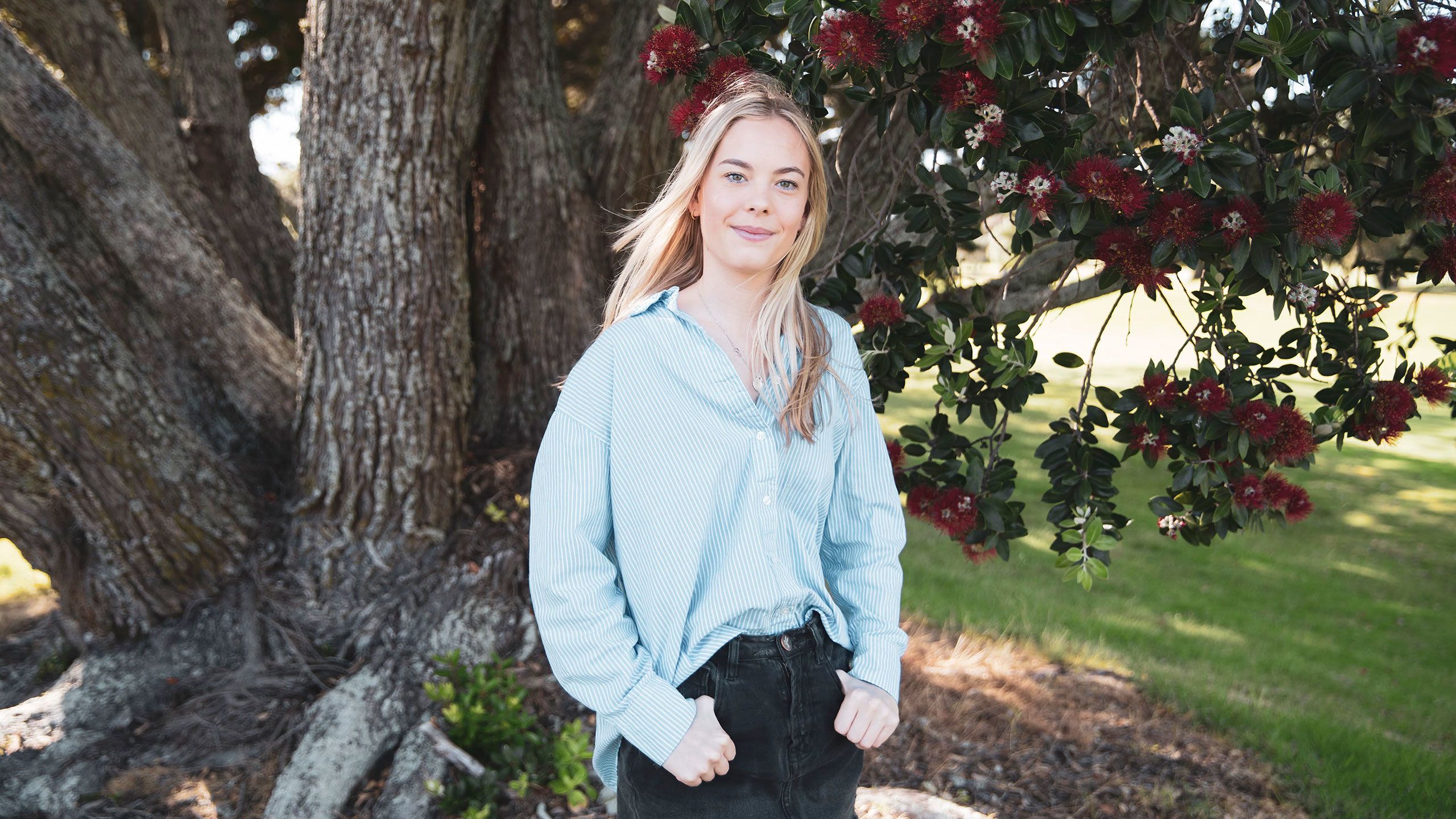 India stands in front of a Pohutukawa tree wearing a pale blue shirt and a denim skirt.