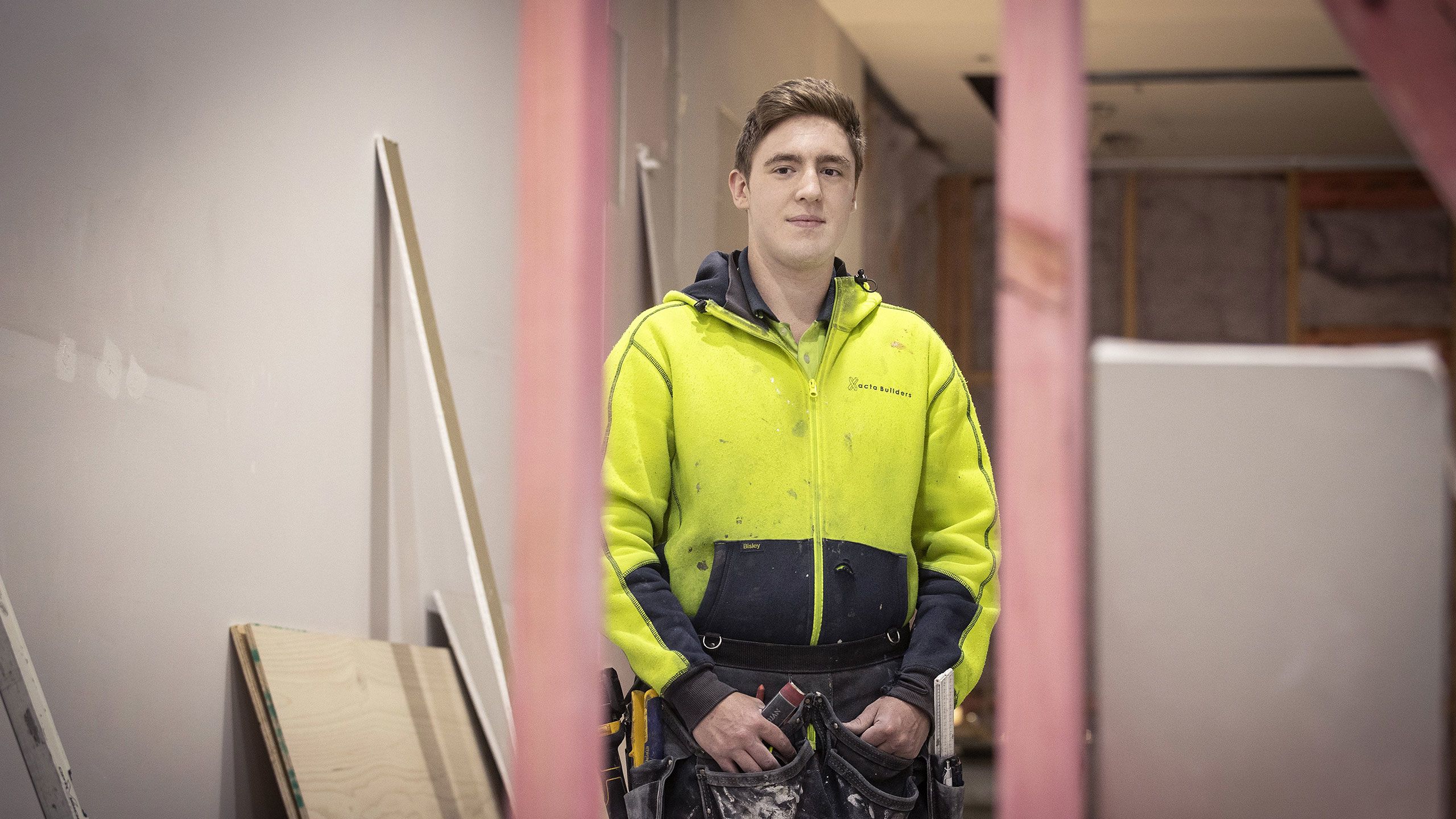 Cameron stands inside a building site in his high-vis sweatshirt. Around his hips he wears his tools in a holster.