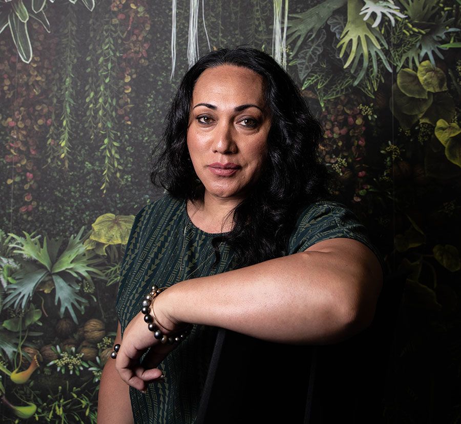 Phylesha Brown-Acton looking directly into camera in front of the tropical forest wallpaper. She has her left arm propped up on the back of a chair.