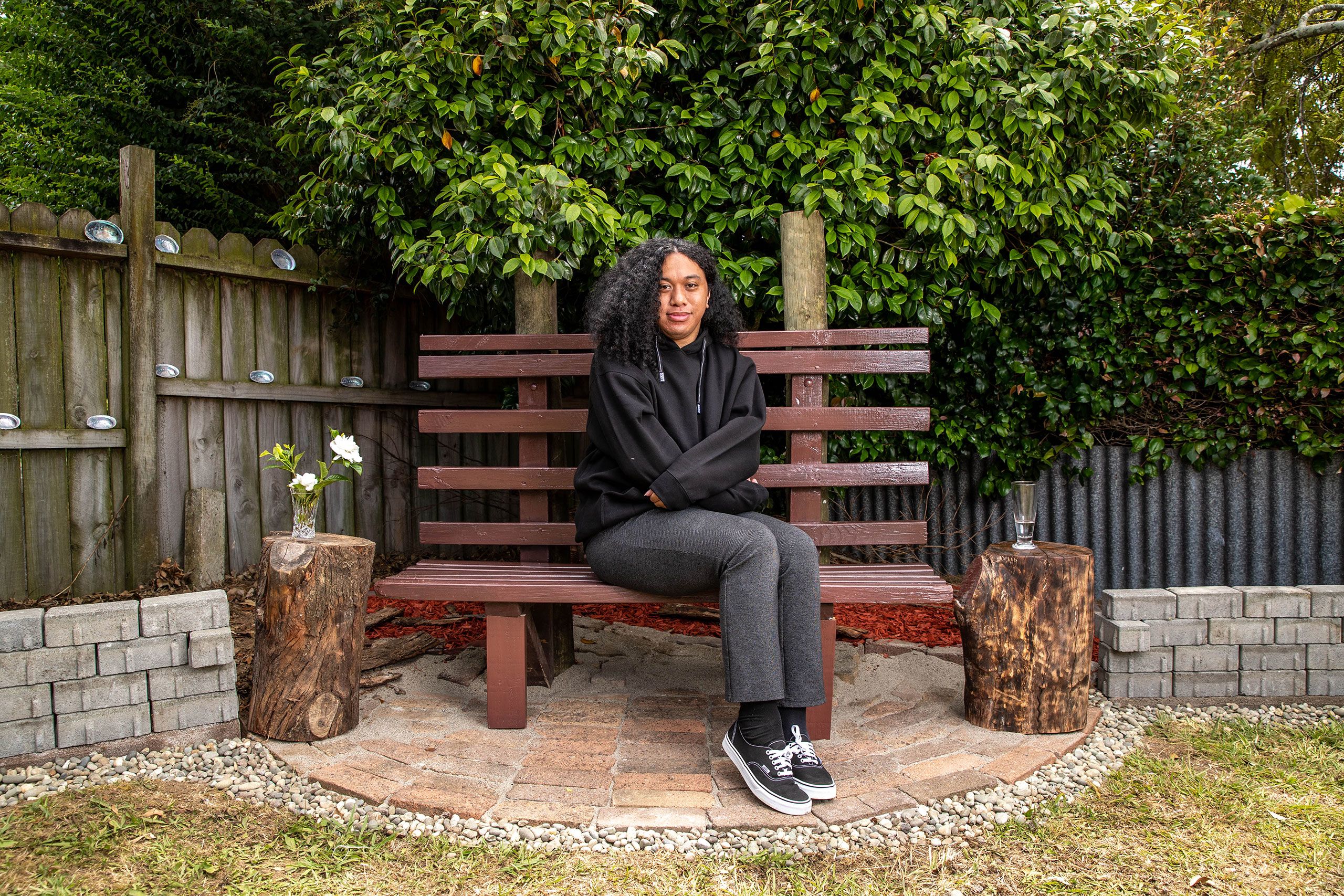 Robin sitting on a wooden bench in the corner of a garden, with a corrugated iron fence and shrubs in the background. She is wearing a black hoodie, grey sweatpants and sneakers.