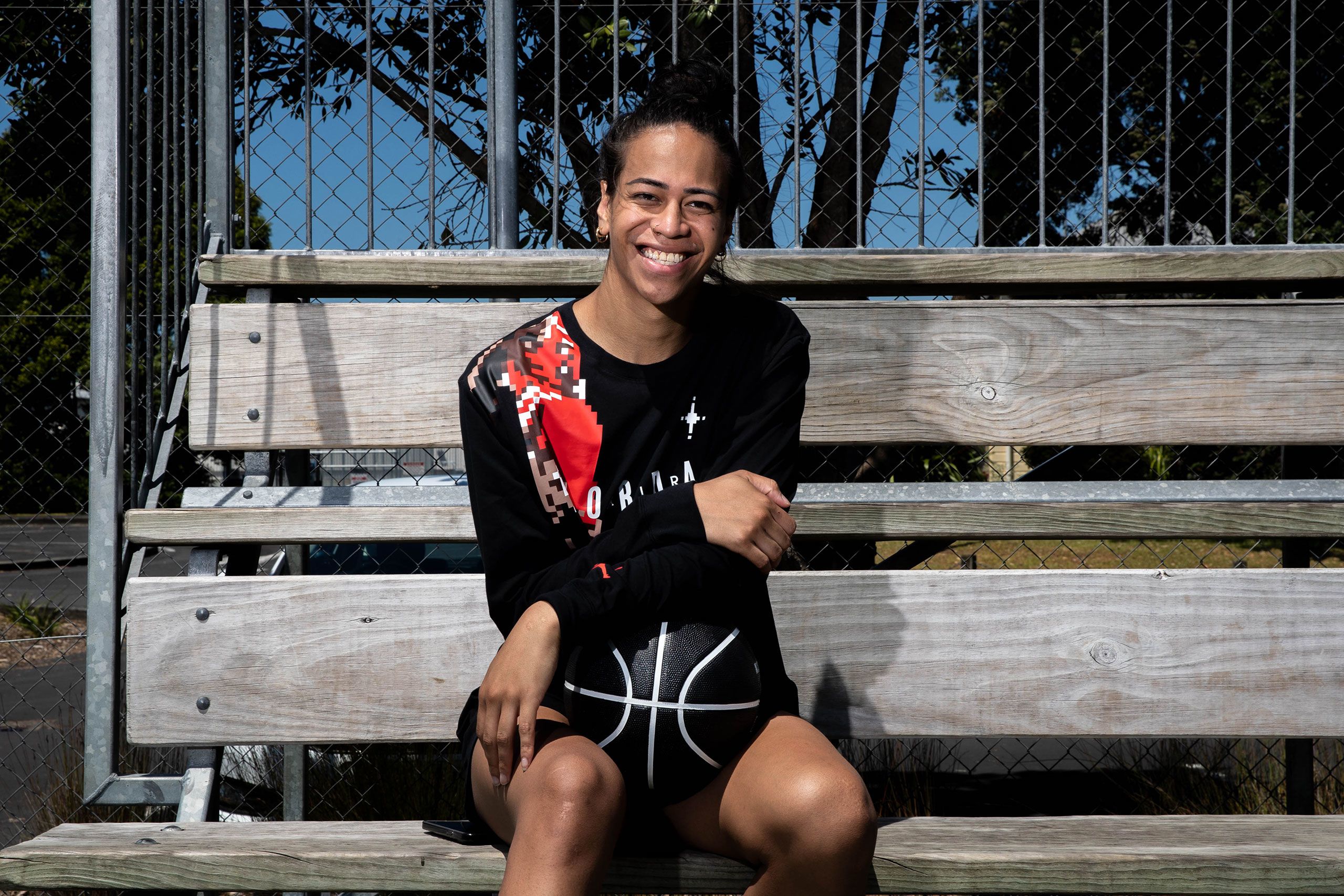 Missy sitting on wooden bleachers, with a chainlink fence, trees and blue sky behind her. She is holding a basketball and smiling directly at the camera.
