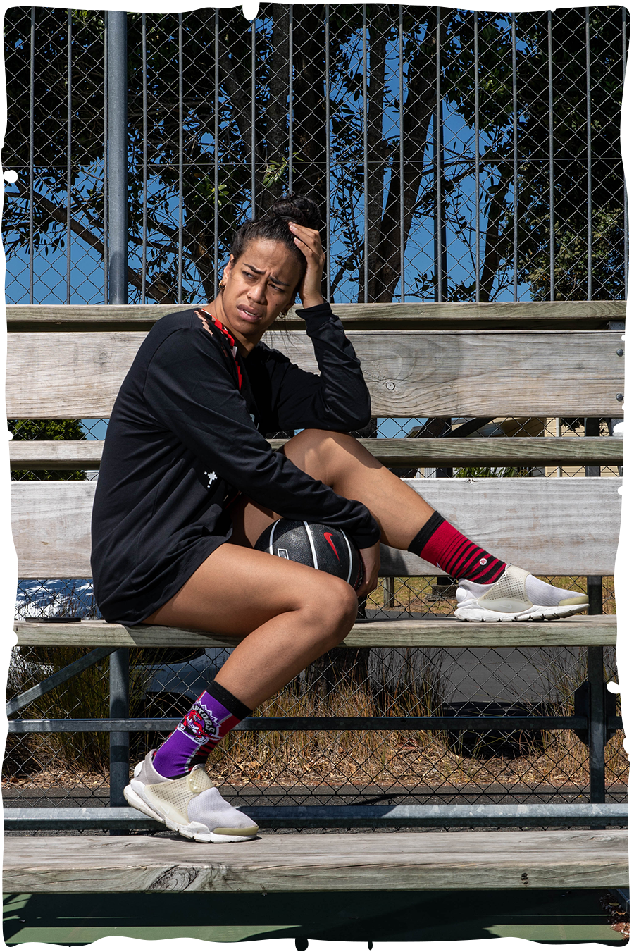 Missy Prescott sitting on wooden bleachers, with her left elbow resting on her propped-up knee. She wears a short black dress, sneakers and red-and-purple socks, and is holding a black basketball.