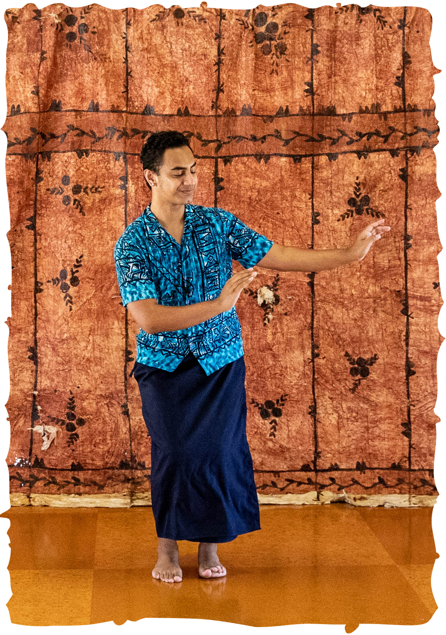 Iatua dancing in front of a large tapa cloth. He is wearing a bright blue patterned shirt and a dark blue lavalava.