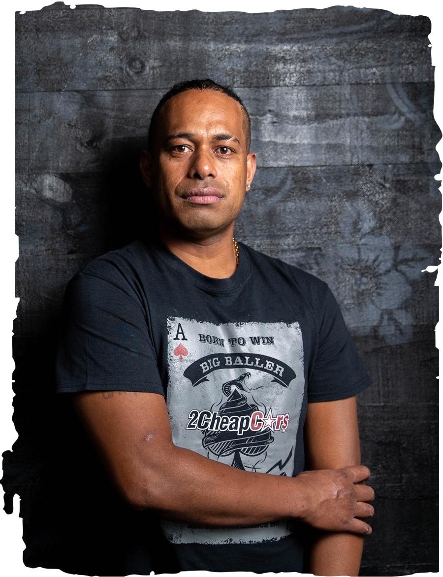 Manasa standing in front of a black wooden wall with a faint hibiscus flower pattern on it. He is wearing a black t-shirt and is looking directly at the camera.