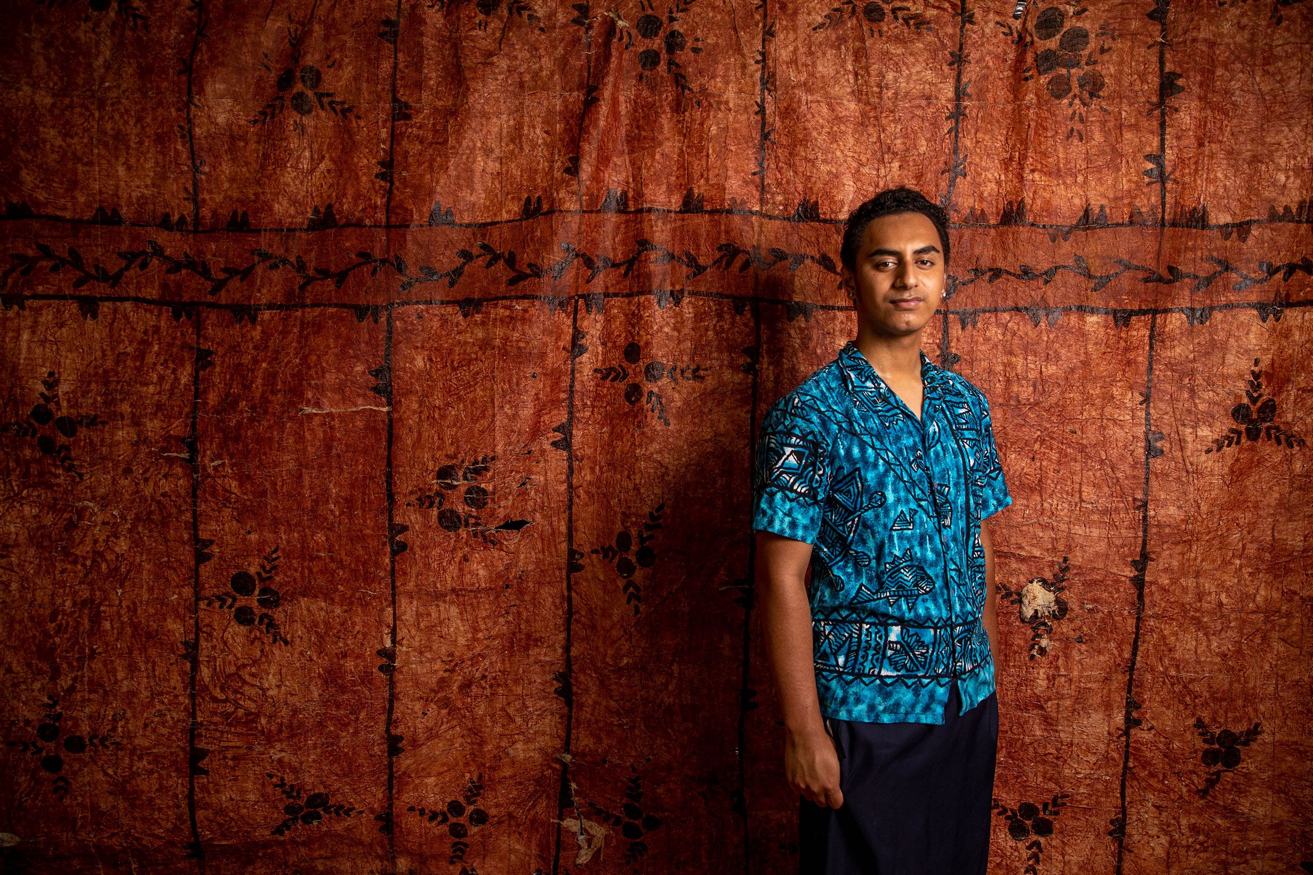 Iatua standing in front of the tapa cloth, looking directly at the camera. He is smiling slightly.